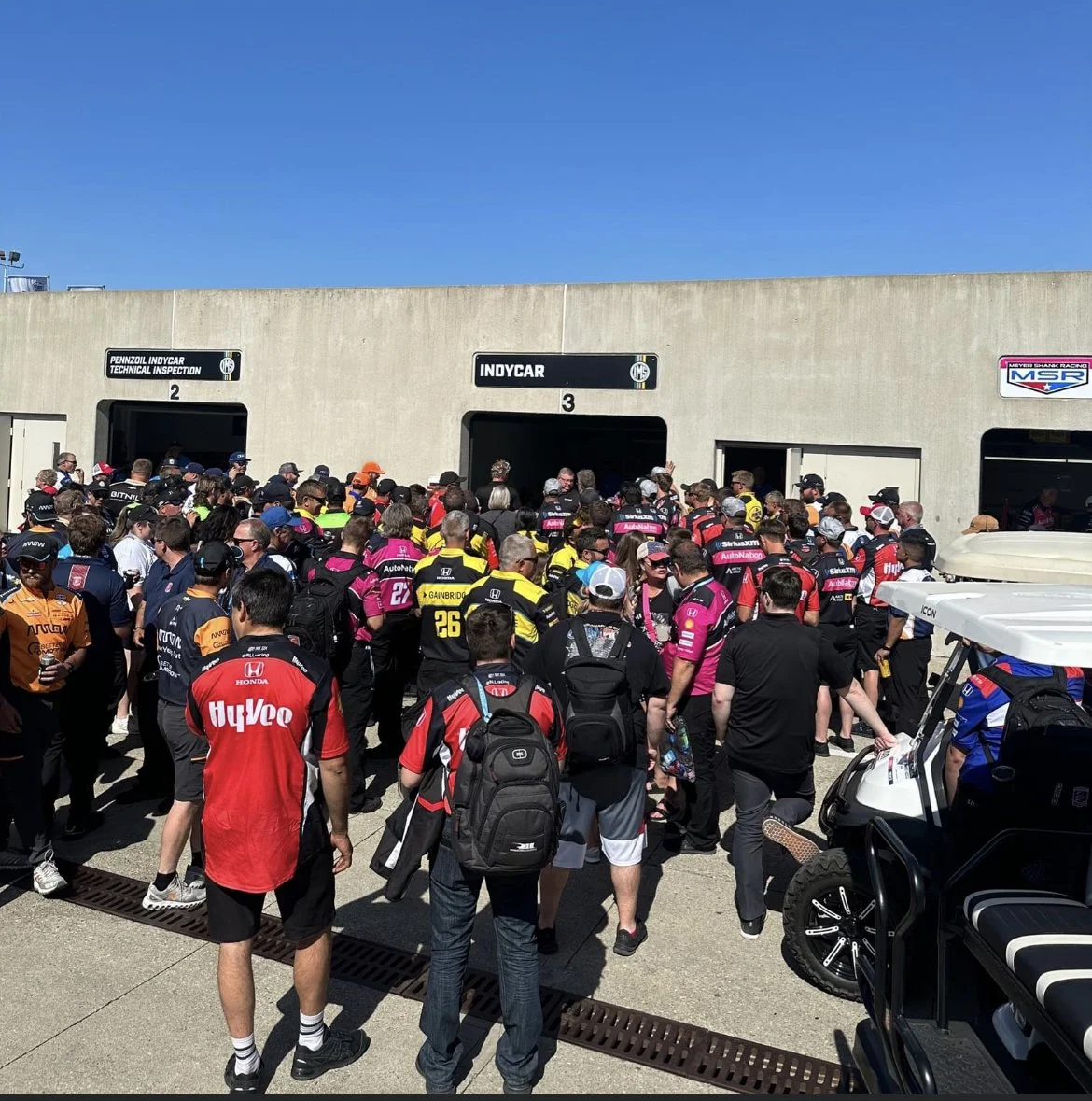 Crowd of race car fans and team members outside Indycar garage at a racetrack on a sunny day.
