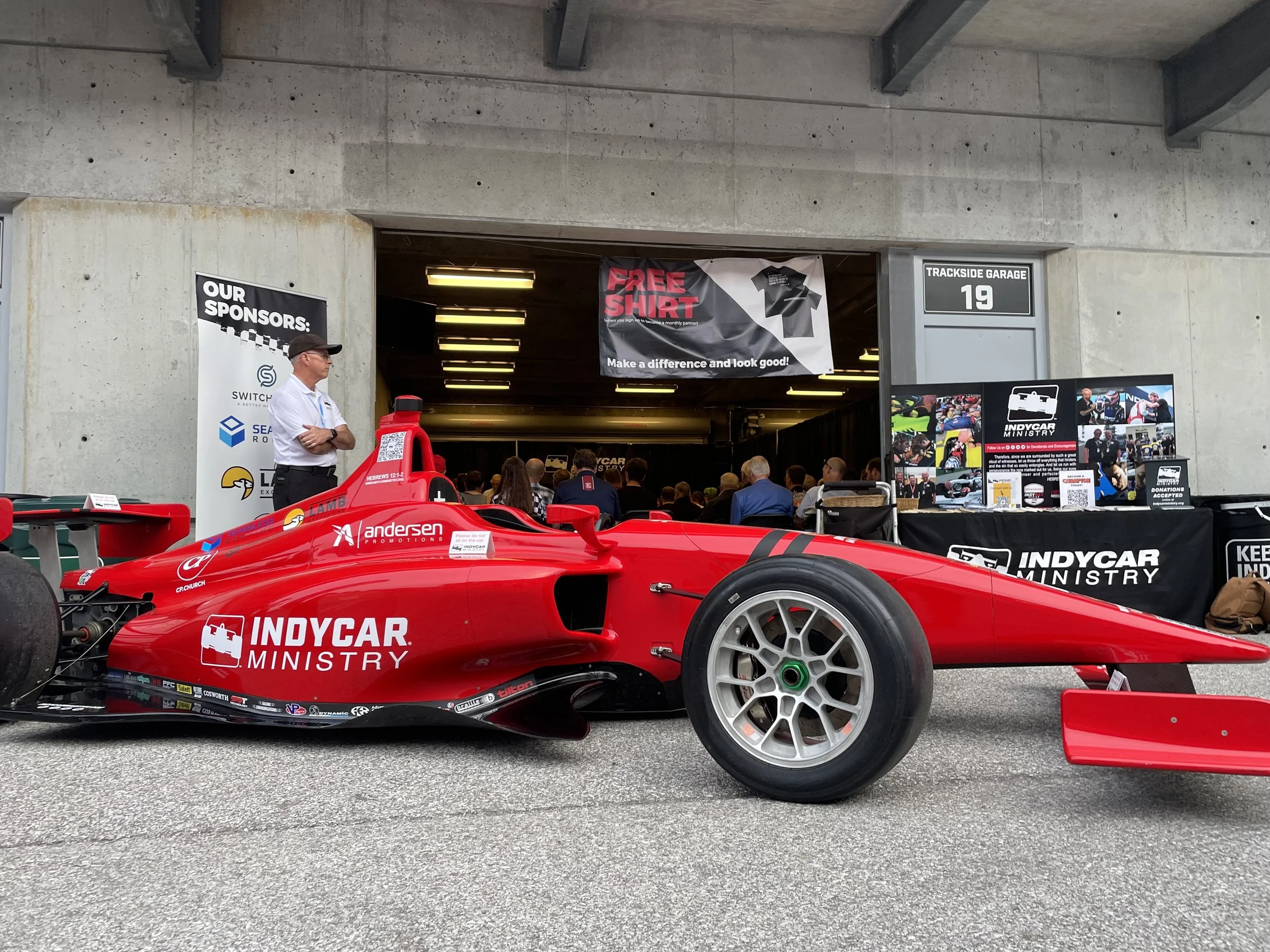 Red IndyCar race car parked outside a garage at a racing event with a man standing nearby and promotional banners in the background.