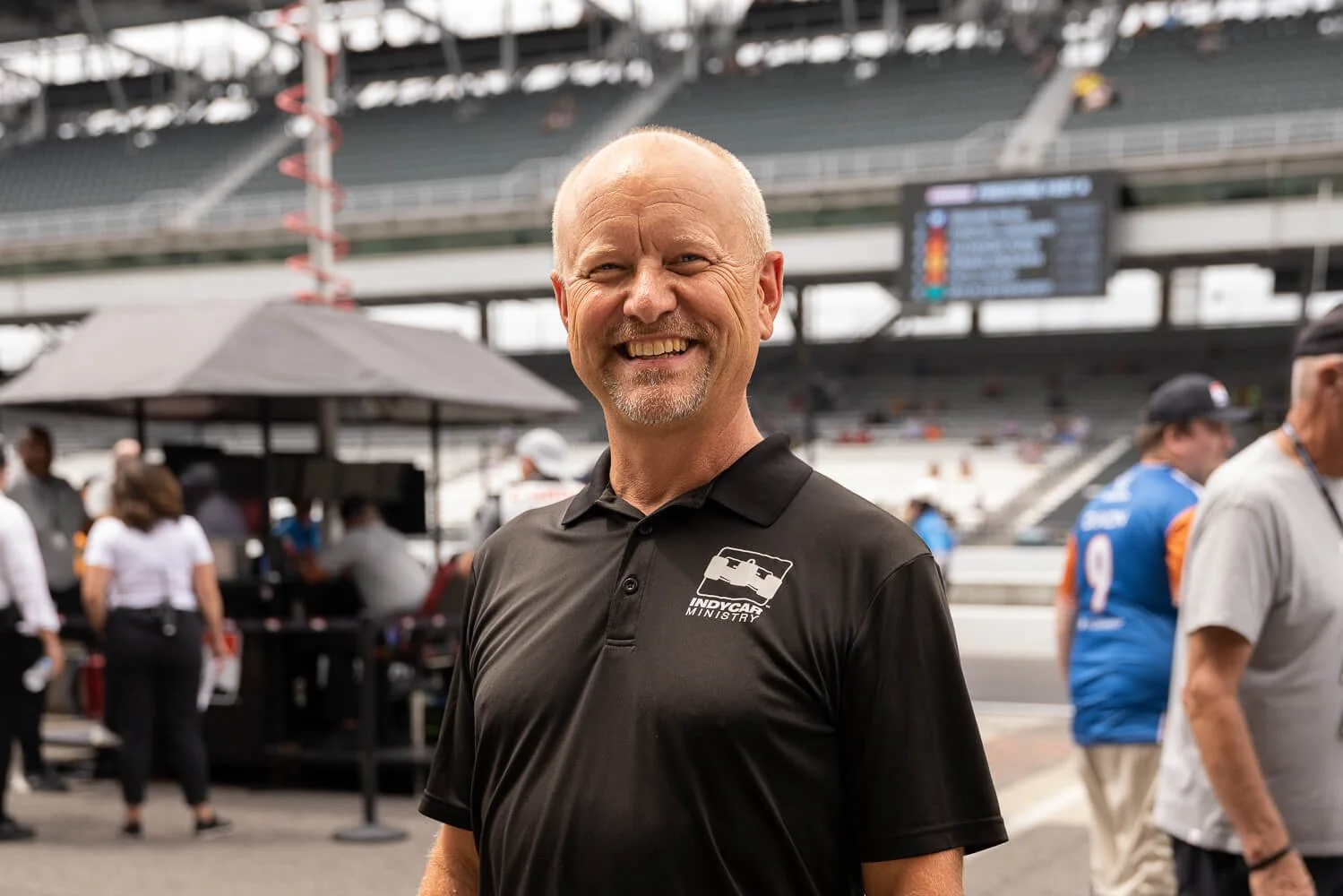 A smiling man wearing a black polo shirt with 'IndyCar Ministry' logo, standing at a race track with a crowd and racing cars in the background.