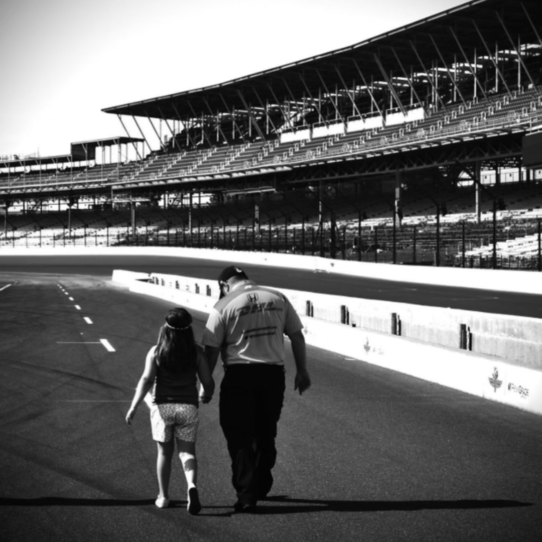 A man and a young girl walking hand in hand on a race track, with empty grandstands in the background.