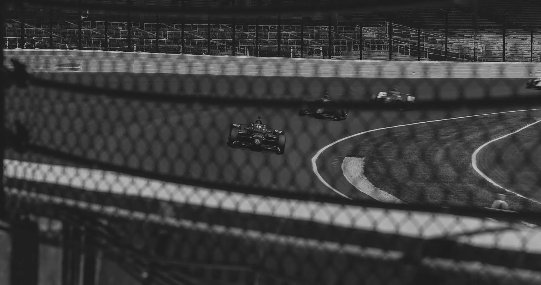 Black and white photograph of race cars on a racing track, viewed through a chain-link fence, with grandstands in the background.