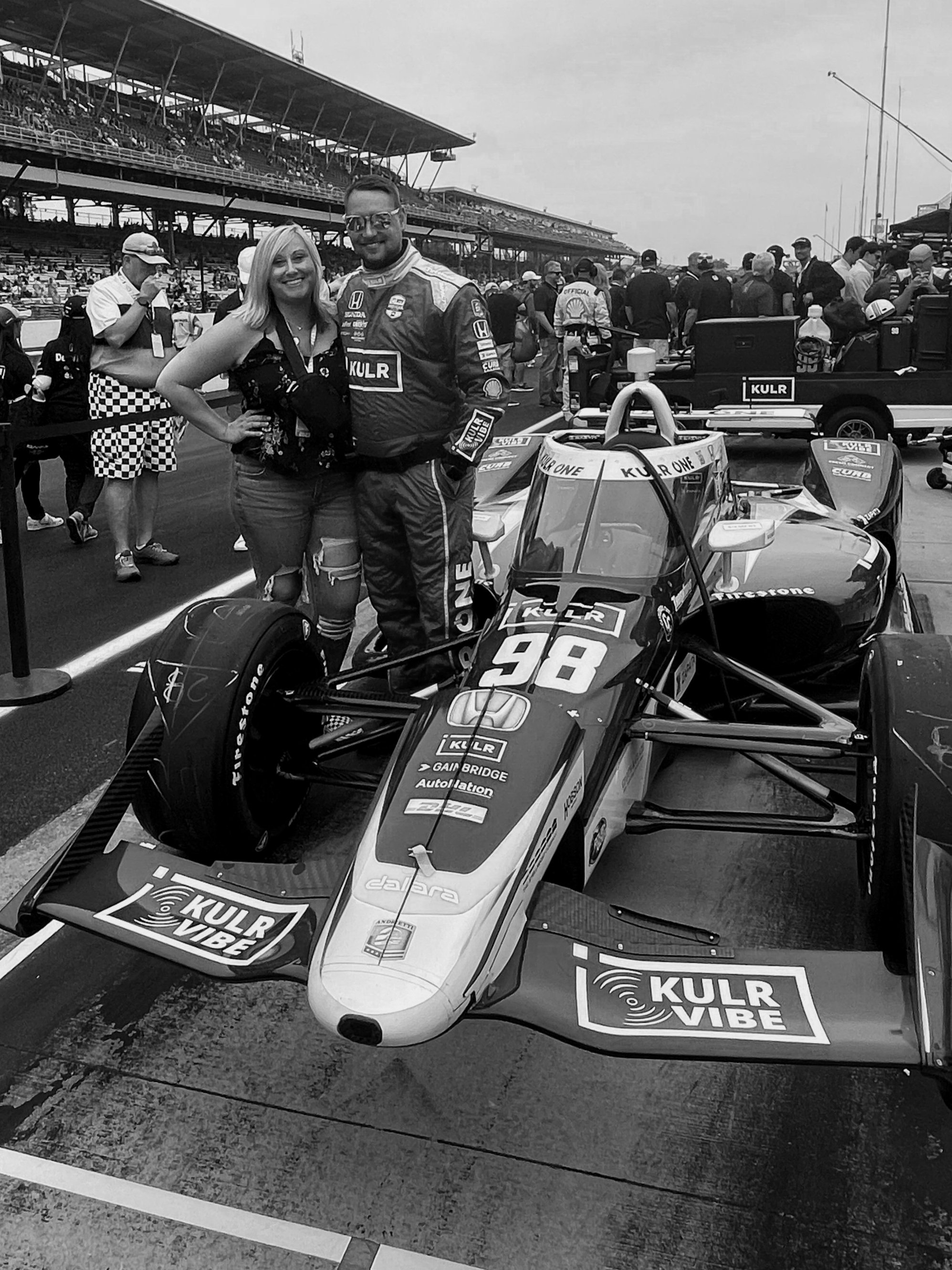 A man in racing gear and a woman are standing next to a race car on a racetrack. There are many people in the background, with some in team uniforms and others watching from the stands.