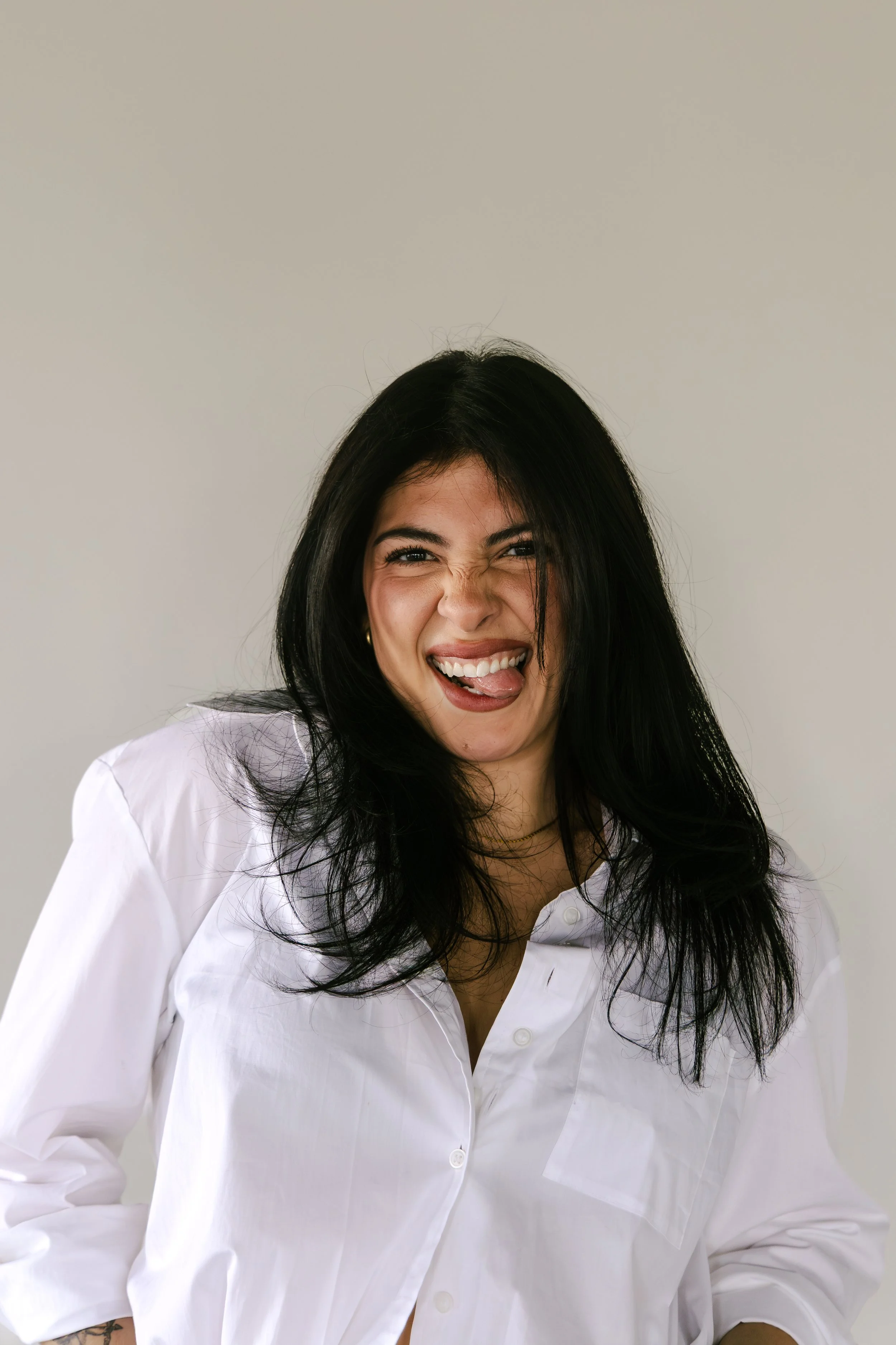 A woman with long dark hair wearing a burgundy off-the-shoulder top and black pants, standing indoors against a neutral background with some leafy plants visible on the side.