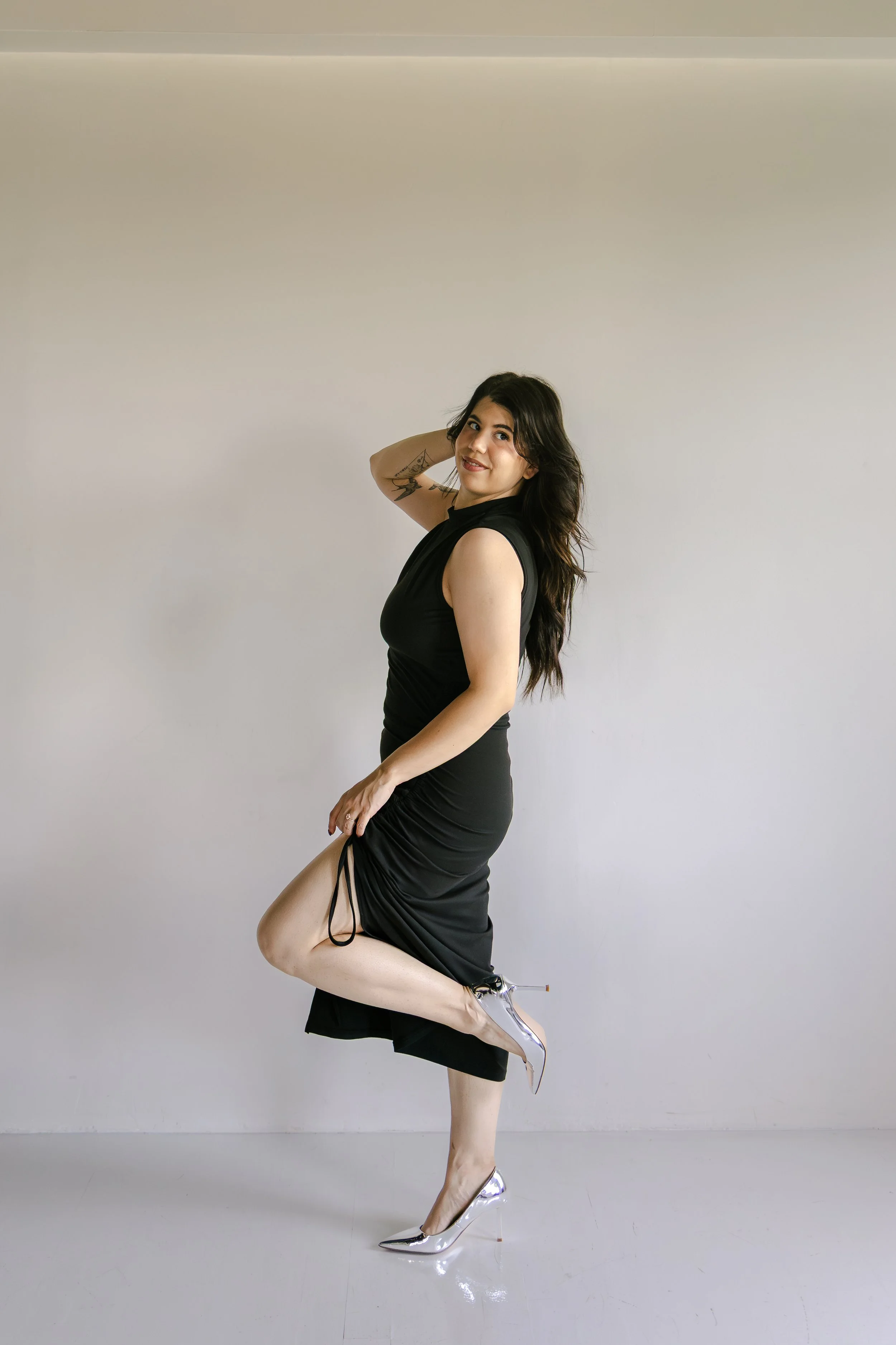 A woman with purple- pink wavy shoulder-length hair, wearing a black sleeveless top with drawstrings on the shoulder and patterned pants, stands in front of a light-colored wall with a plant branch on the right.