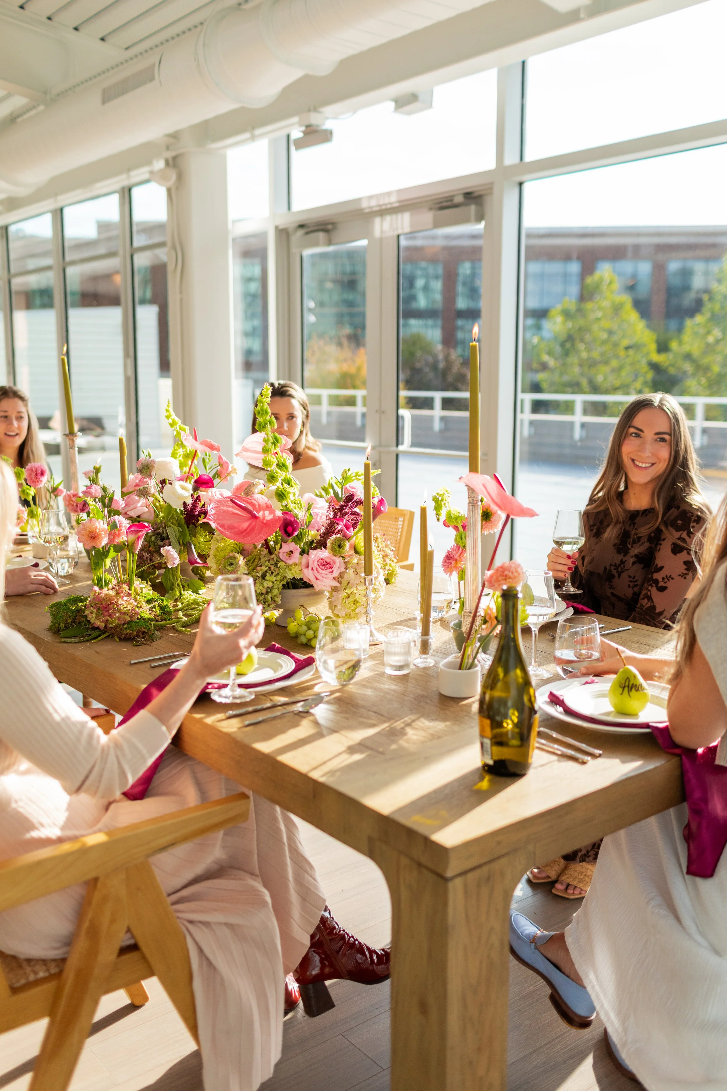 Dining table decorated with pink and white flowers, candles, and a personalized pear place card, with a person holding a glass of white wine.