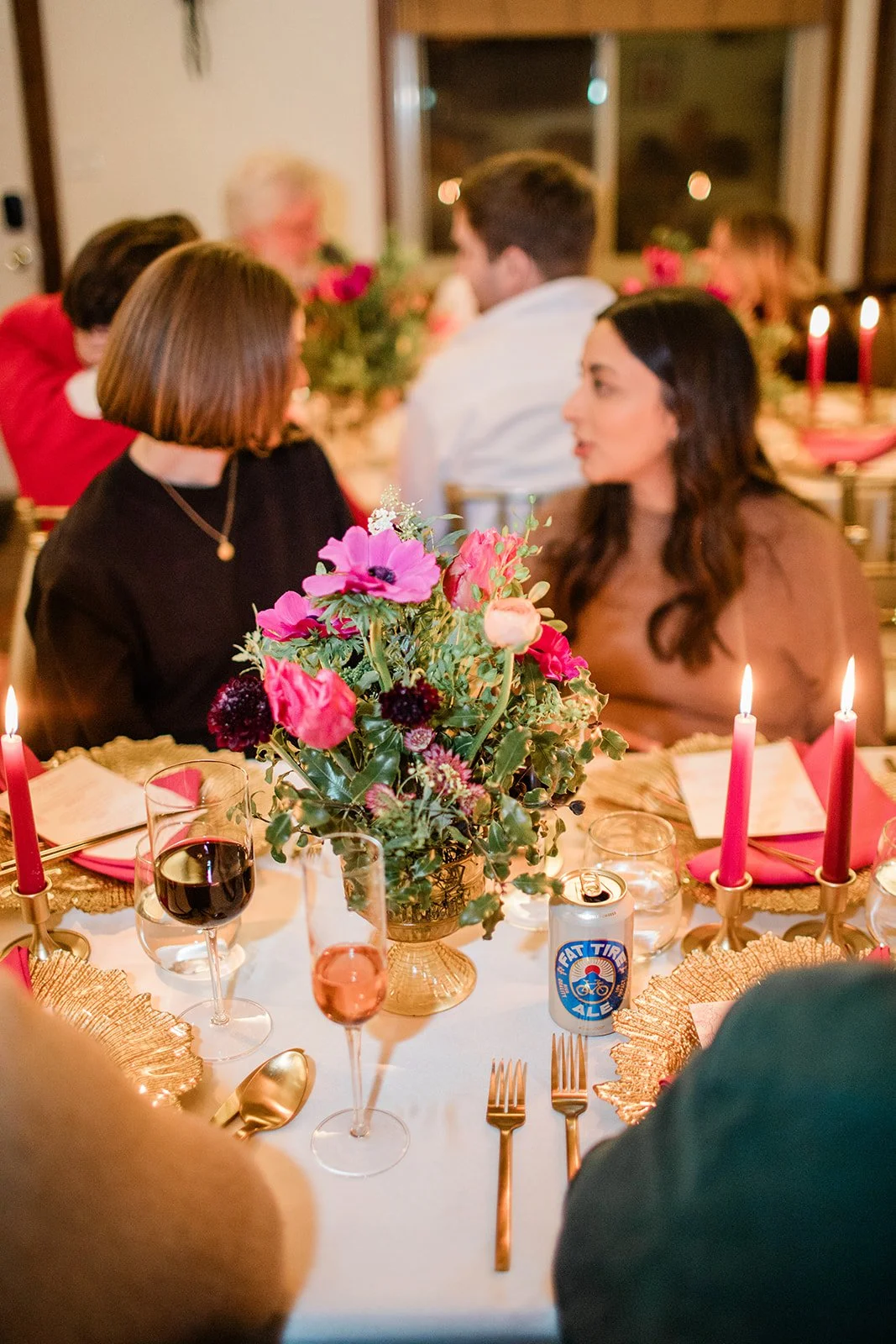 A festive dinner table decorated with a large colorful flower centerpiece, pink and red candles, pink napkins, gold cutlery, glasses, and a can of beer. People are seated around the table, engaged in conversation, with some blurred in the background.