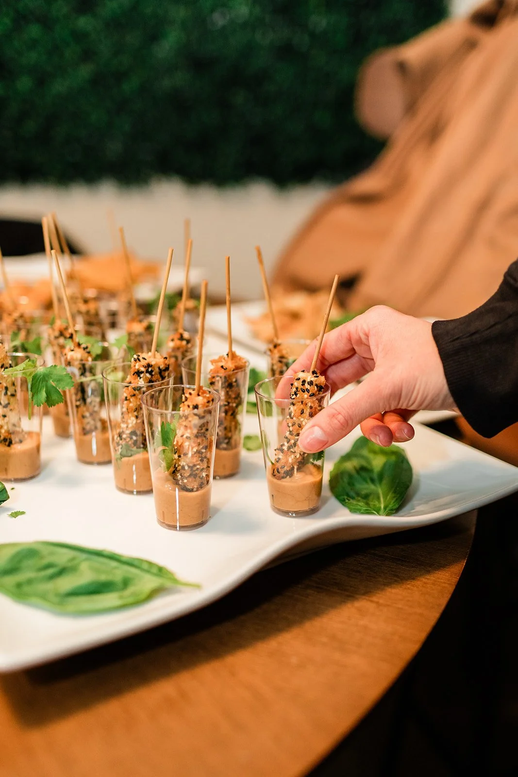 Person placing a sesame-crusted appetizer with a toothpick into a shot glass filled with creamy sauce on a white serving platter decorated with lettuce leaves.