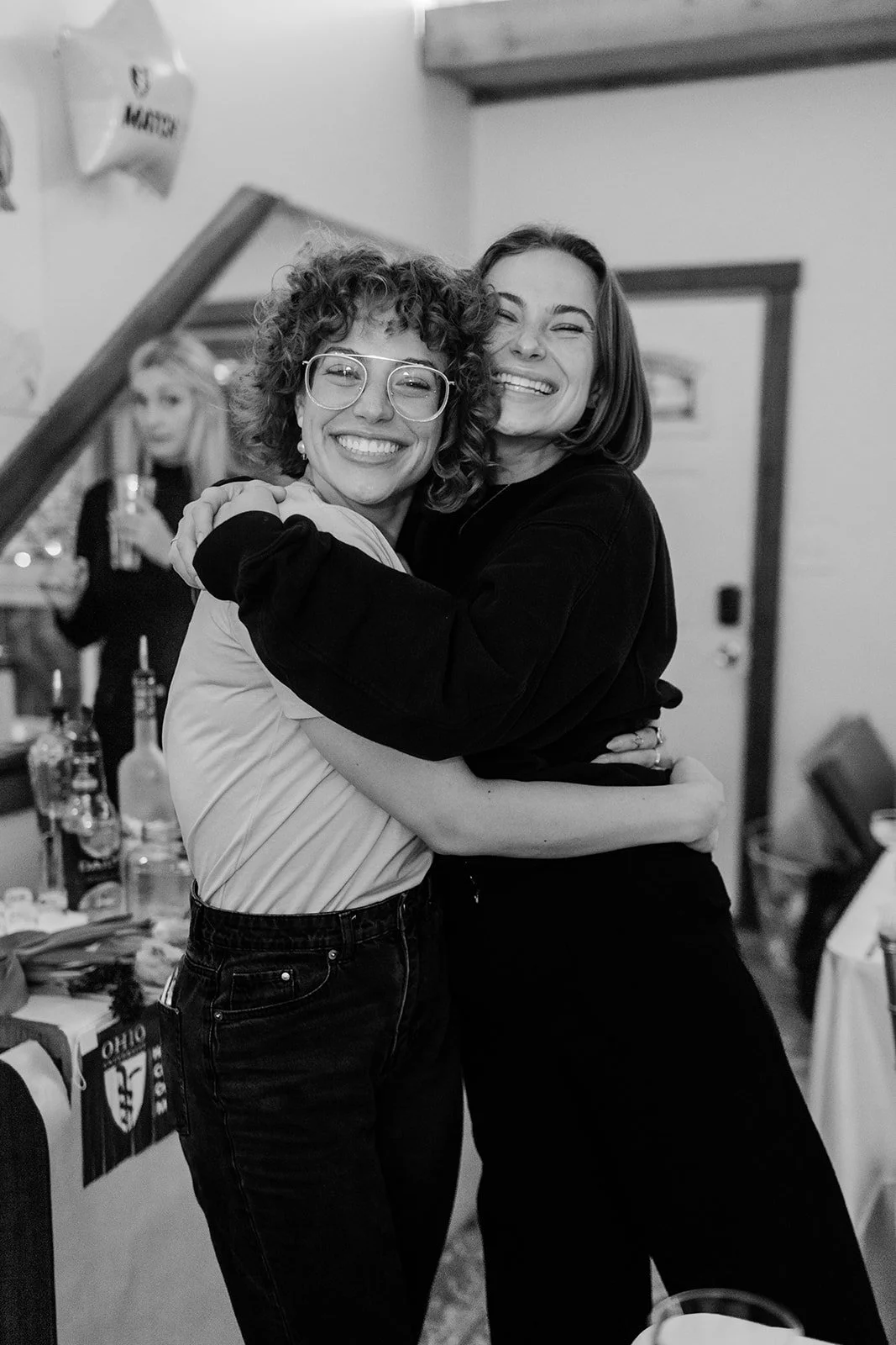 Two women smiling and hugging each other at a social gathering, with a woman in the background holding a drink.