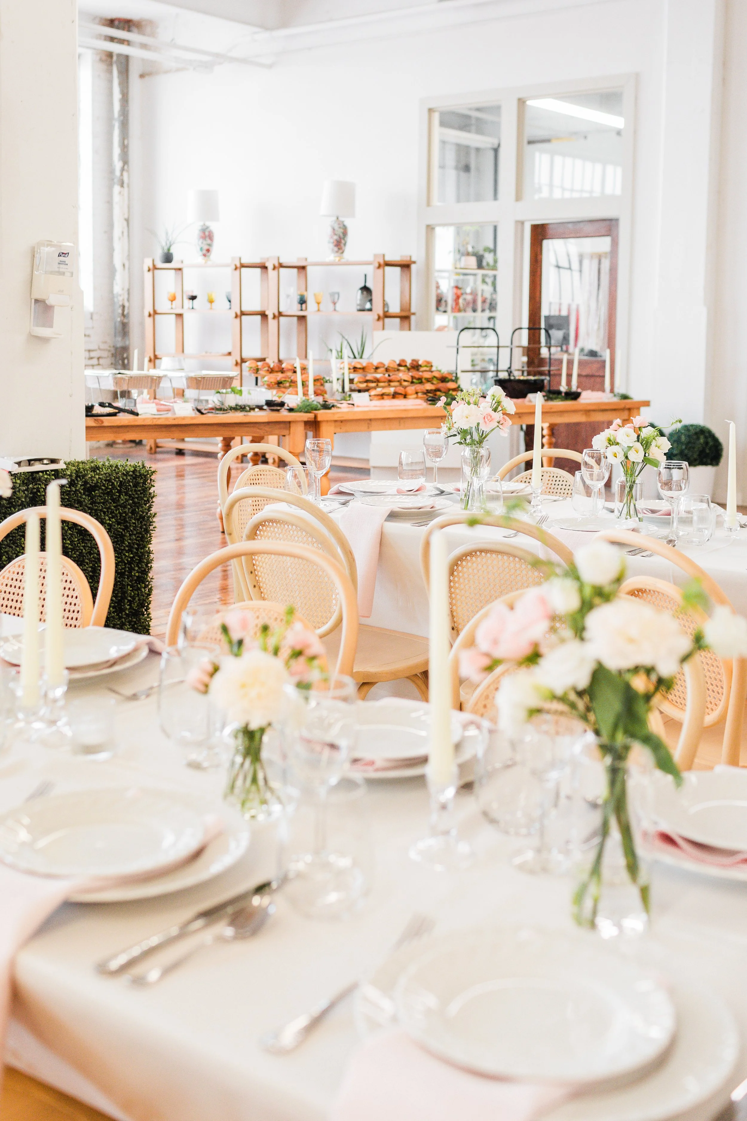 Elegant dining setup with round tables, floral centerpieces, candles, and neatly arranged plates and cutlery, for a celebration or special event in a bright, spacious room.