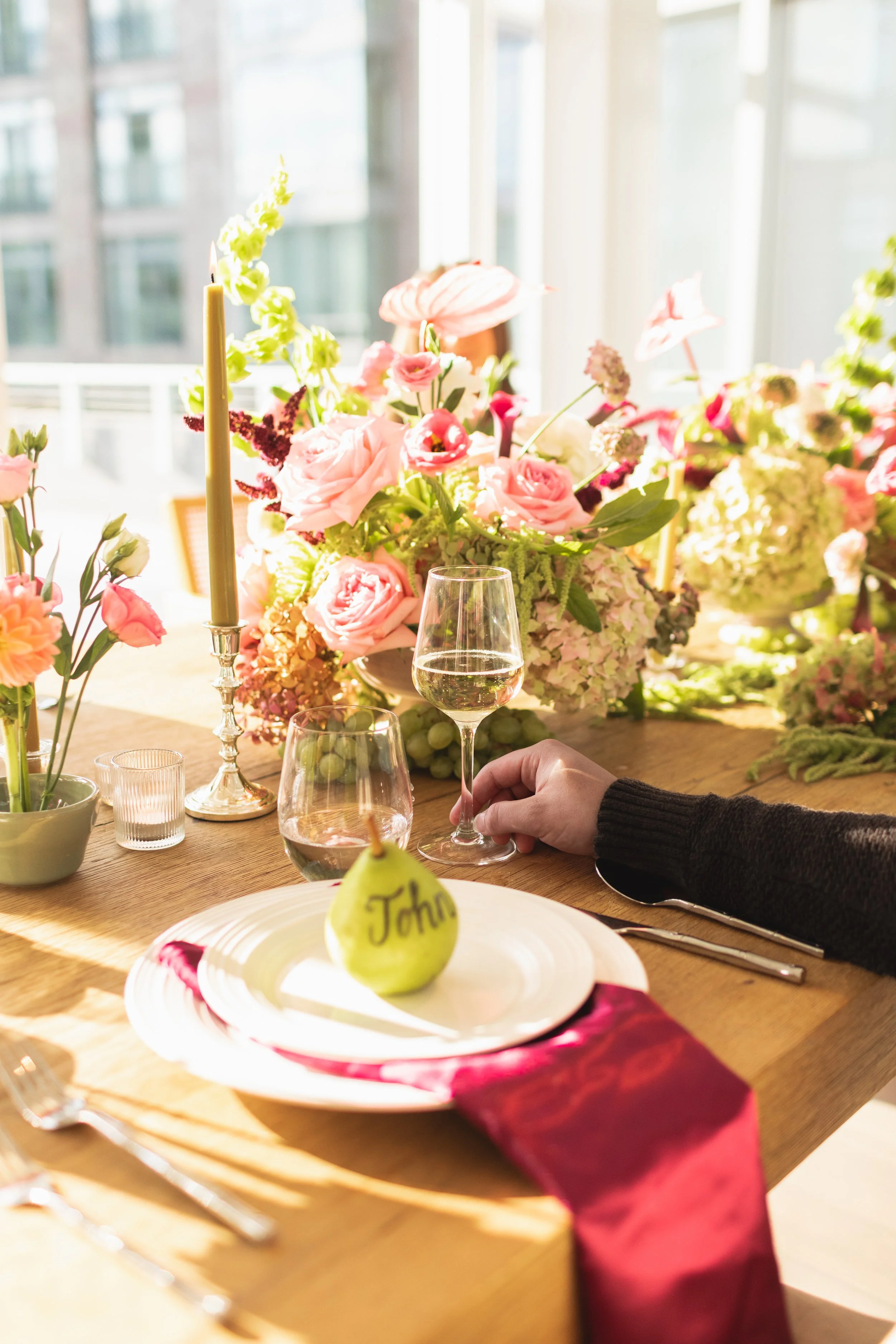 A dining table set with pink flower arrangements, a pear with the name 'John' written on it, wine glasses, a candlestick, and utensils, with sunlight coming through large windows.