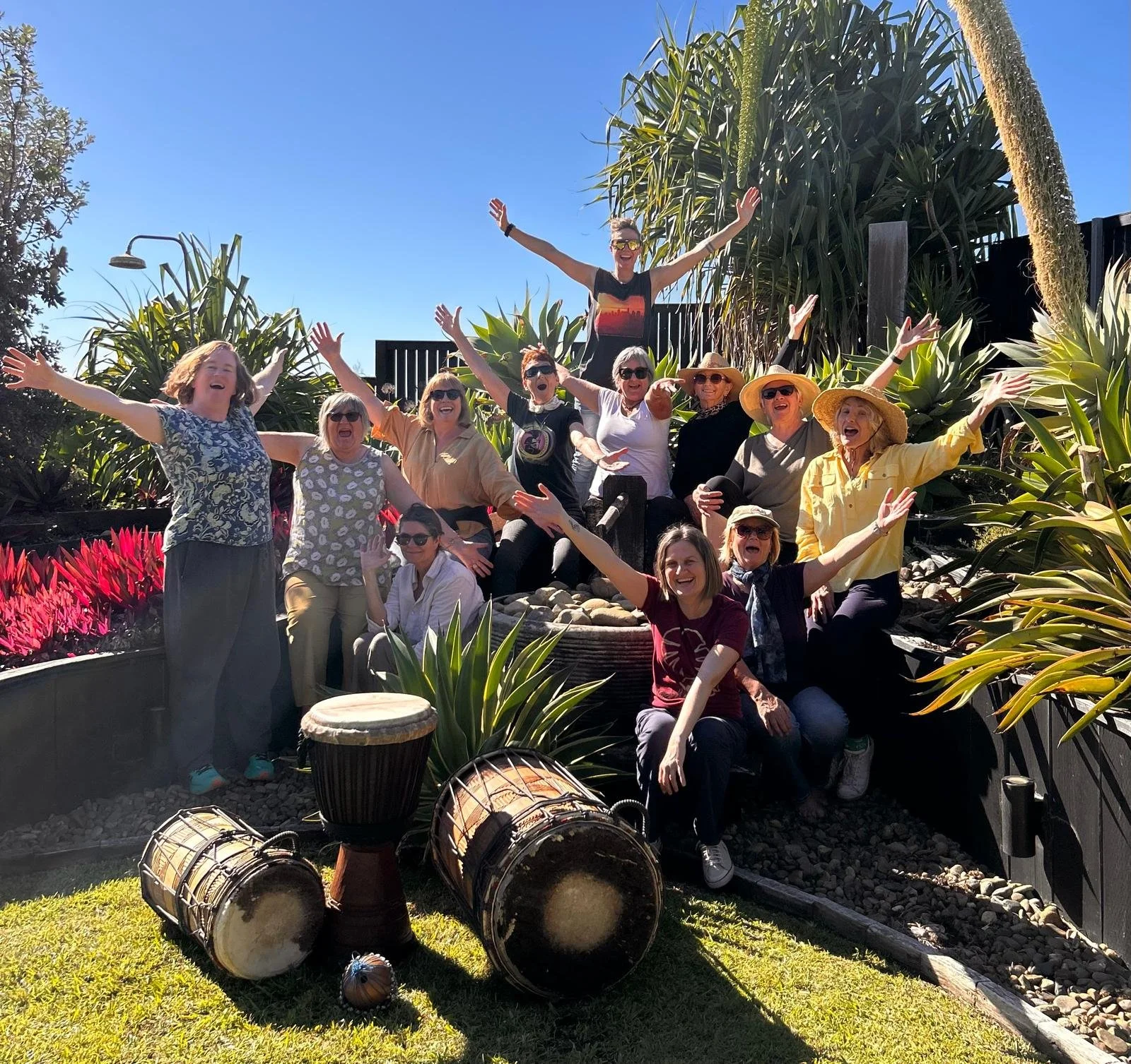 A group of people smiling and celebrating outdoors in a garden with lush green plants, drums, and a clear blue sky.