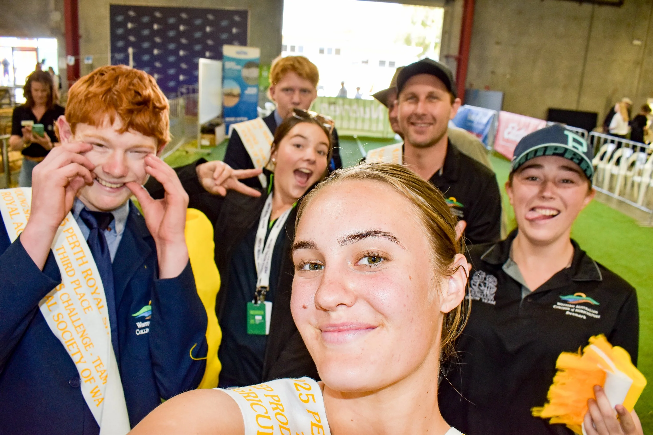 Group of six people taking a selfie at an indoor event, smiling and making playful gestures, with some holding yellow and white cloths, and wearing medals around their necks.