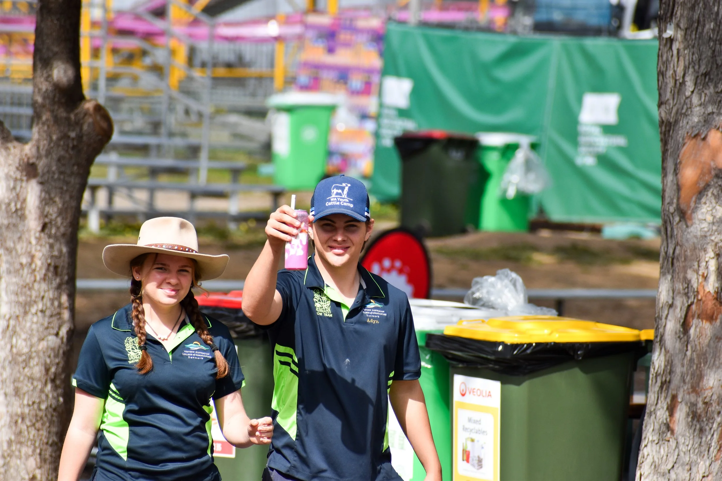 Two young people, a girl with braids wearing a wide-brimmed hat and a boy with a cap, smiling and walking outdoors near trash bins, holding a pink drink.