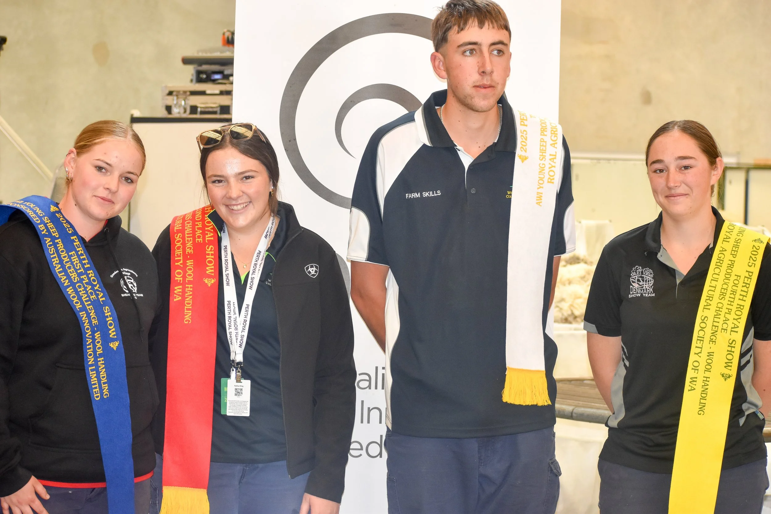 Four young people at an awards ceremony, wearing medals and sashes, with a backdrop displaying the event details. Three females and one male, smiling and posing together.