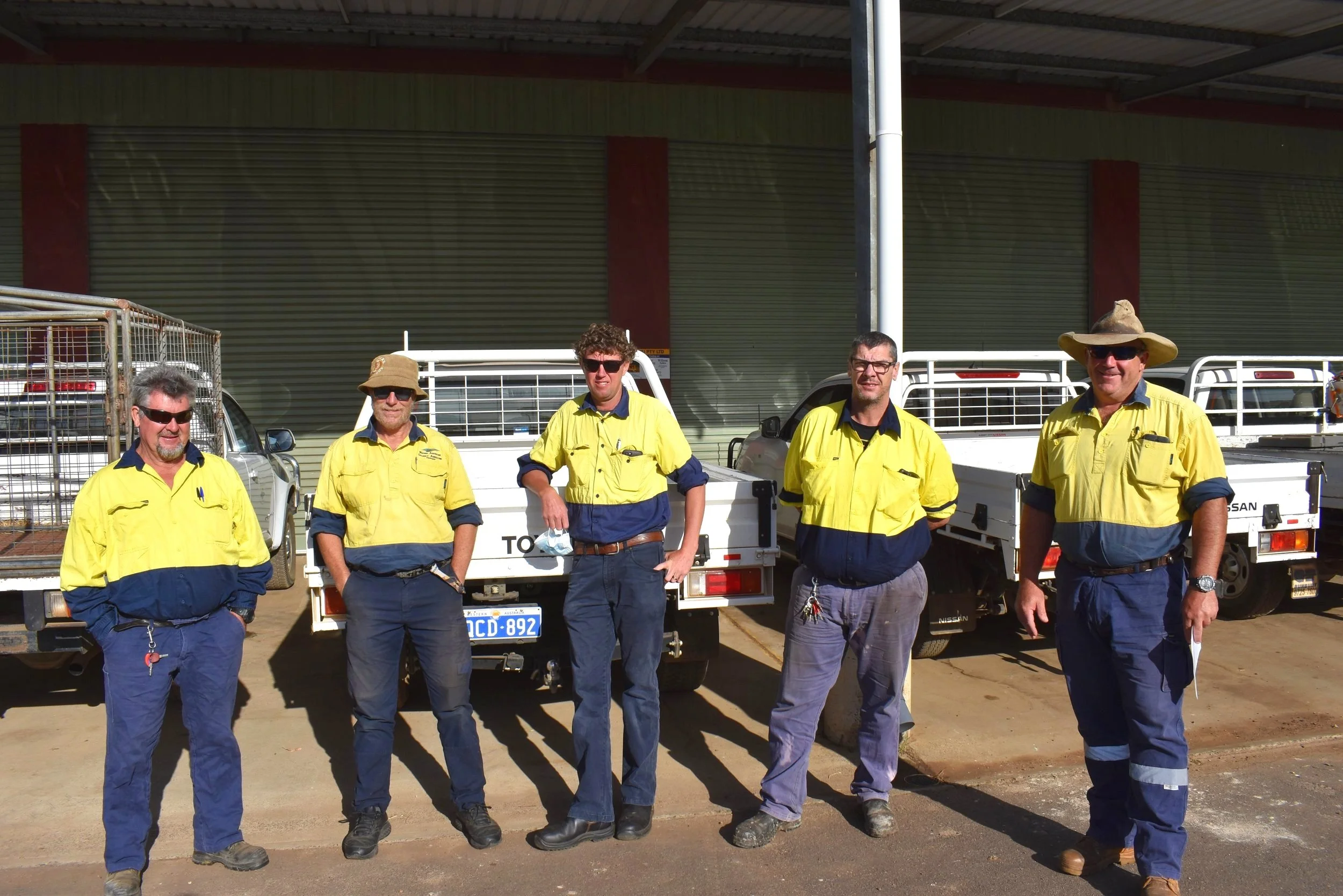 Five men in yellow and navy uniforms standing outdoors in front of trucks.