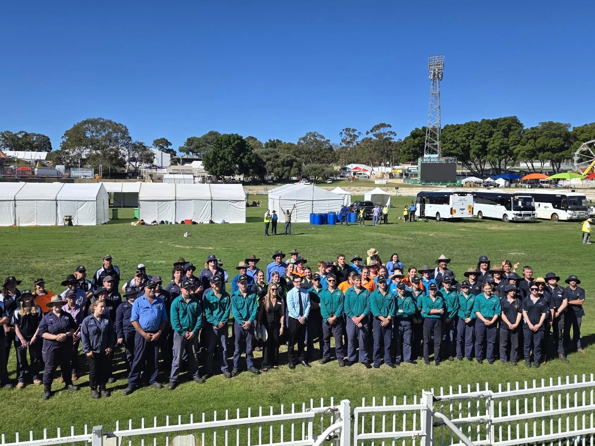 Group of people, some wearing uniforms and hats, standing on a grass field at an outdoor event with tents, buses, and trees in the background.