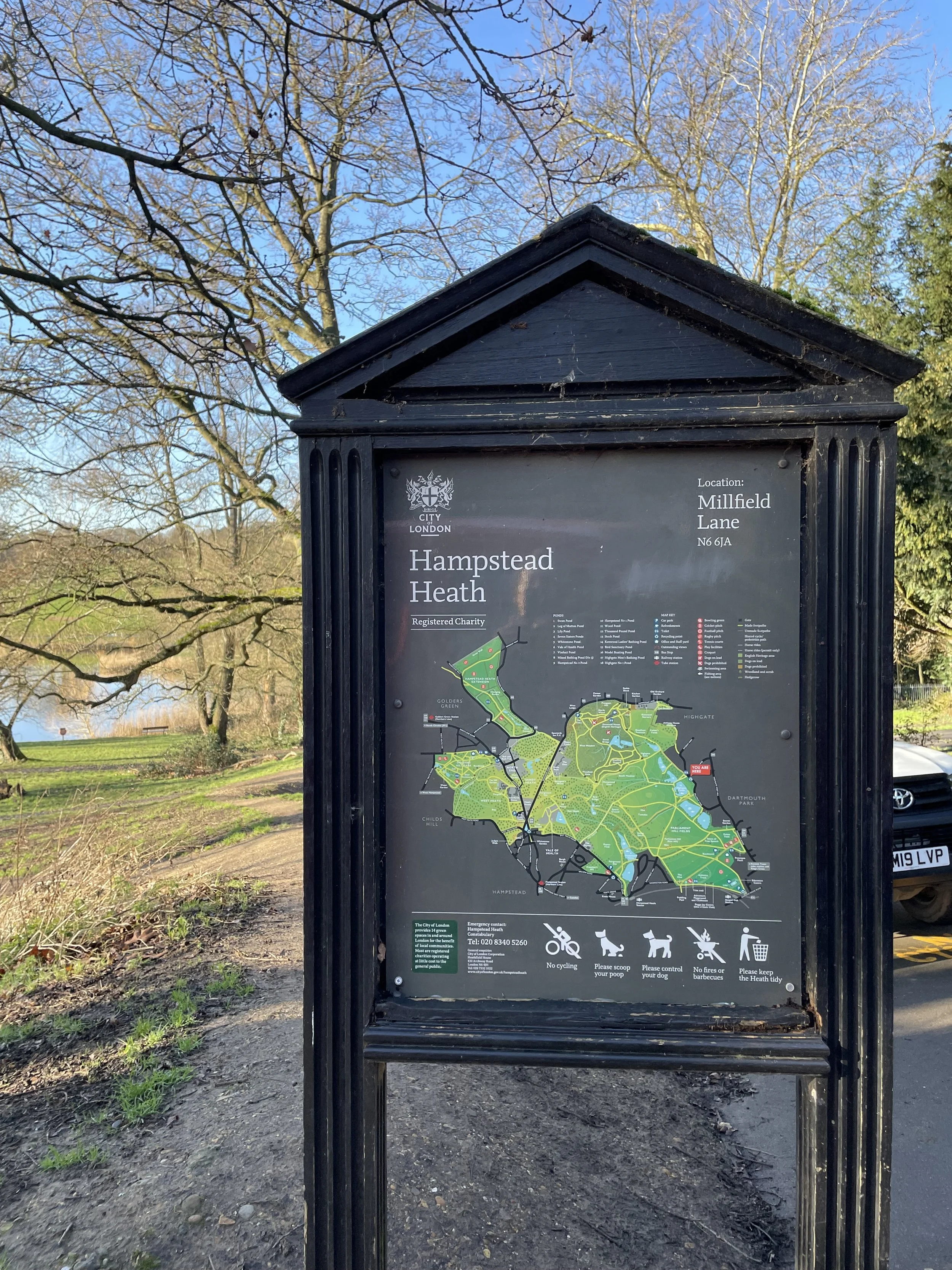 Sign and map of Hampstead Heath in Millfield Lane, London, surrounded by trees and parked car.
