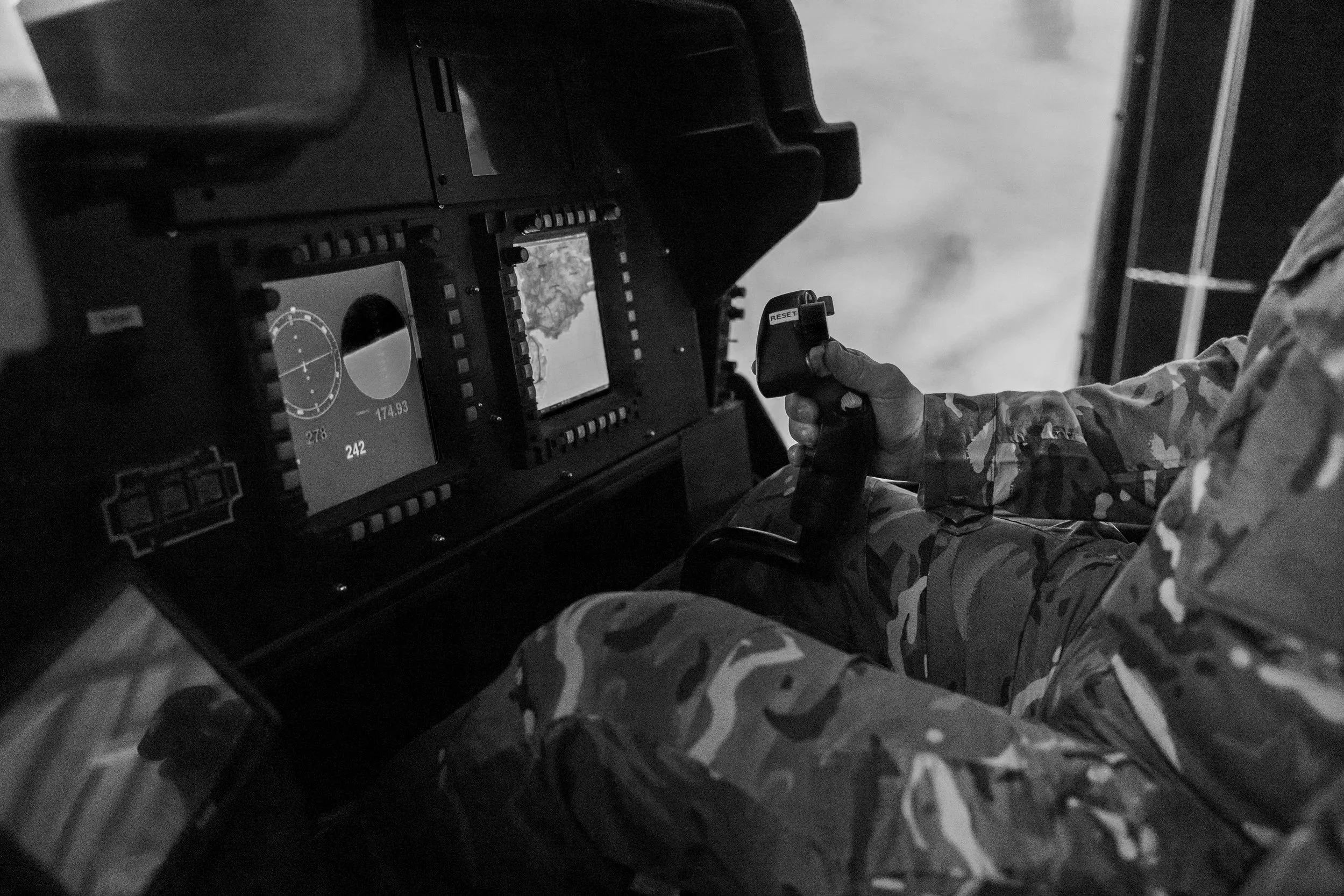 A person in military camouflage uniform sitting inside a cockpit with two digital screens, one displaying a radar and the other a map, controlling with a joystick.