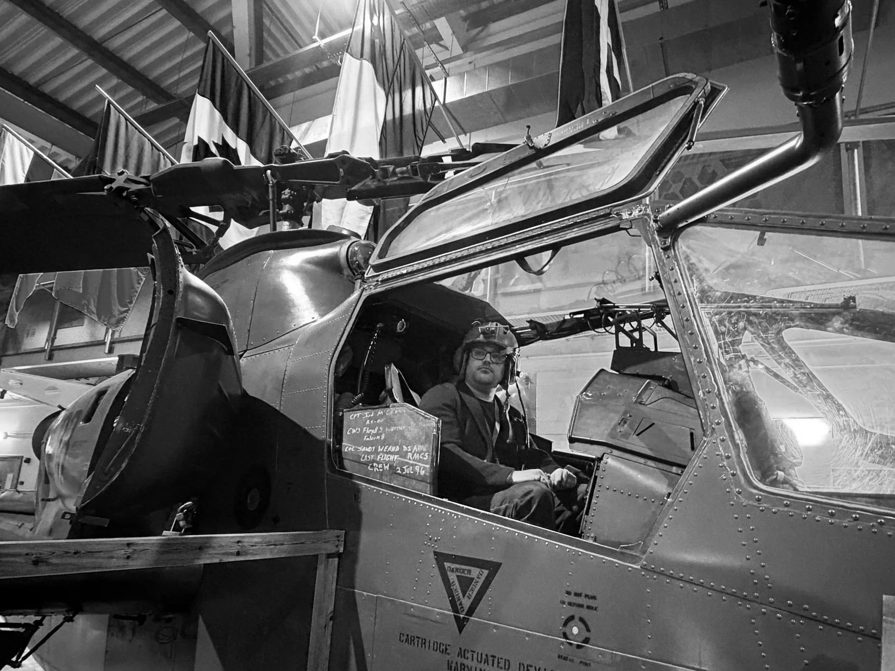 A person wearing glasses and a pilot's helmet sitting inside a vintage military aircraft cockpit, with flags hanging in the background.