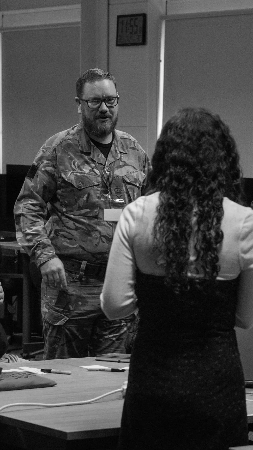 A man in military camouflage speaking to a woman with curly hair at a table, in a room with a digital clock showing 11:55 on the wall.