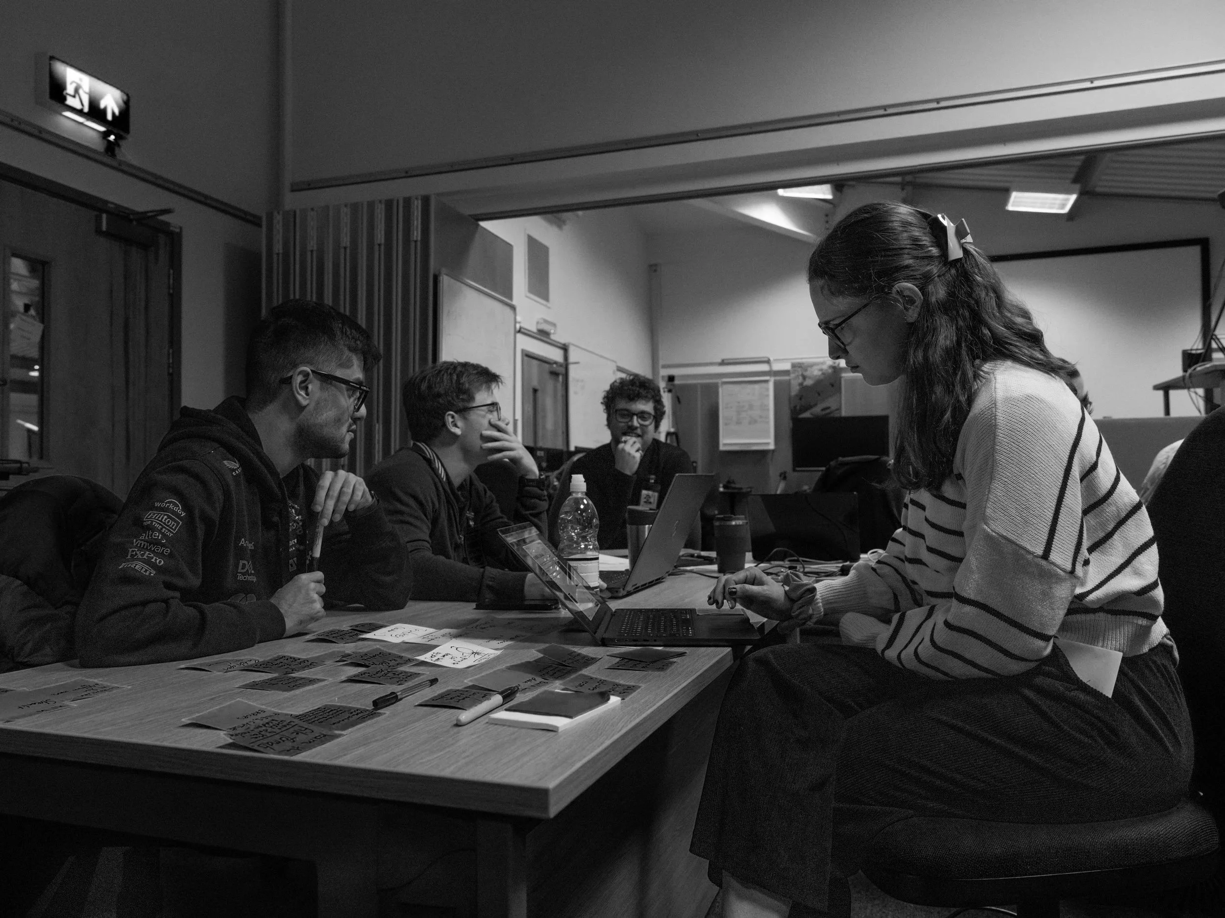 Four people sitting at a conference table with paper notes, a laptop, and water bottles, engaging in discussion in a room with a whiteboard and a projector.