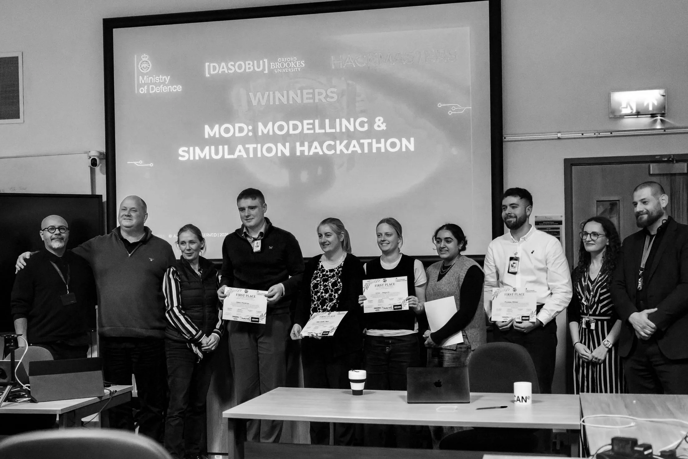 Group of nine people standing in front of a presentation screen at a hackathon event, some holding certificates, celebrating winners of the Modelling & Simulation Hackathon.