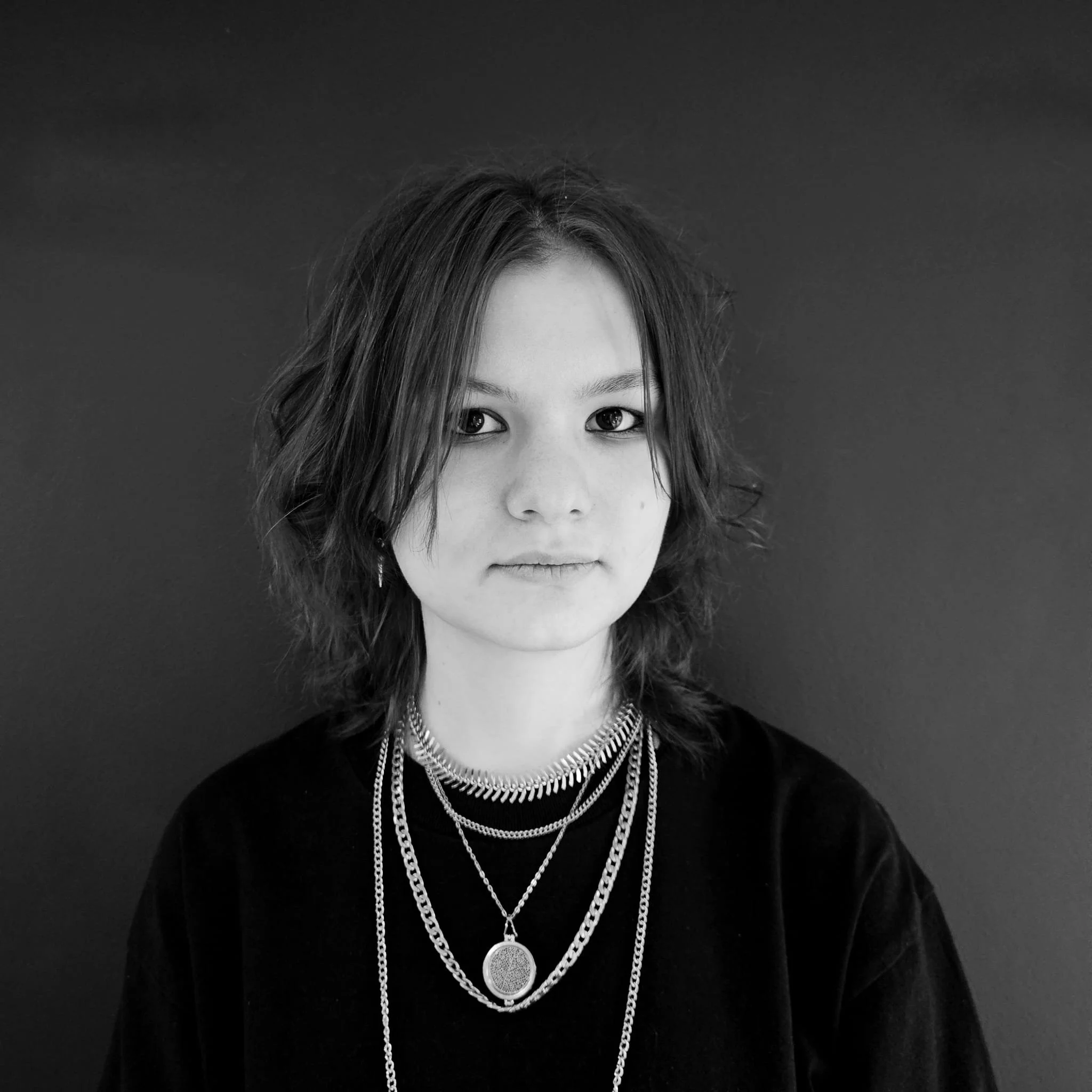 Black and white portrait of a young woman with shoulder-length wavy hair, wearing layered necklaces and a dark top, standing against a plain dark background.