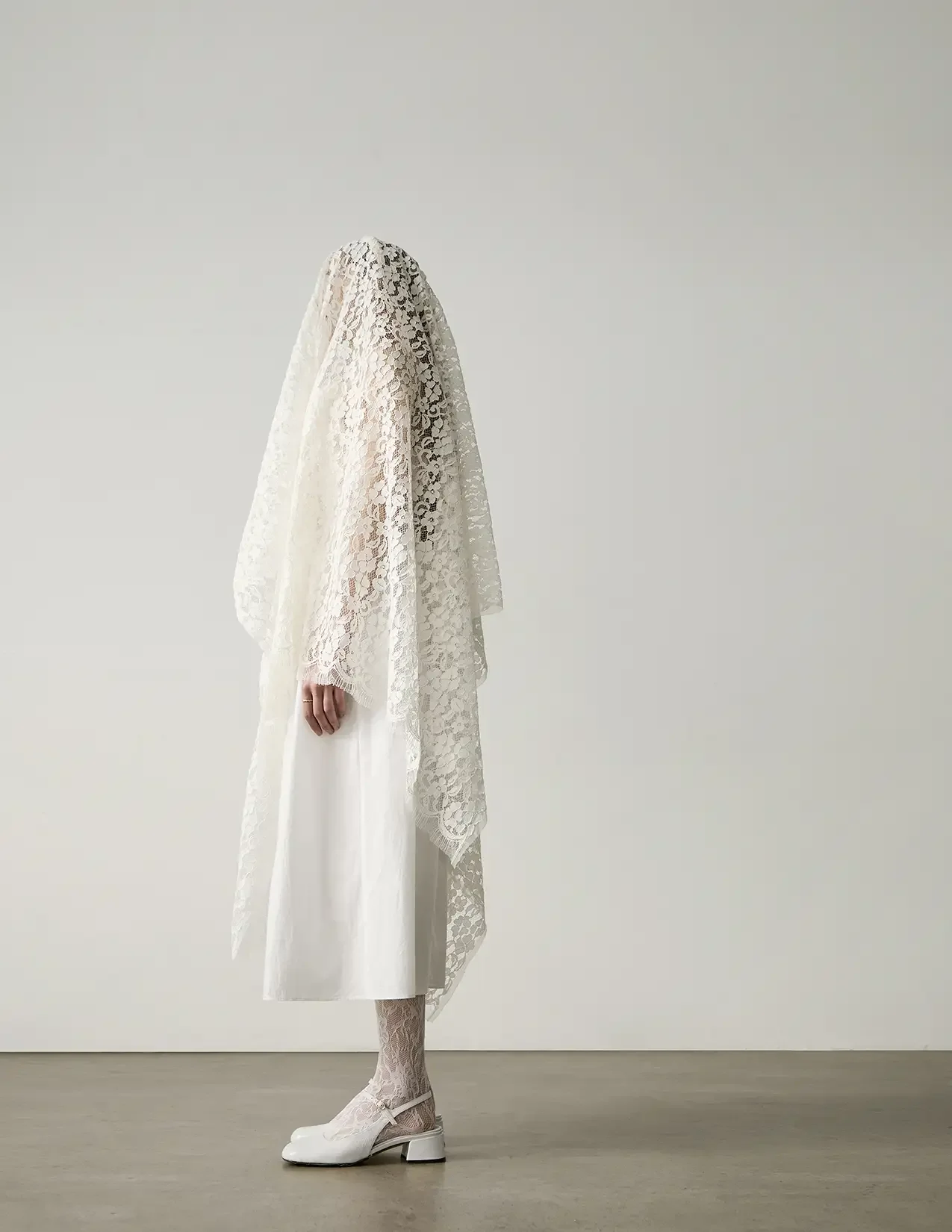 A person wearing a white lace garment, with lace shoes and stockings, standing against a plain background.
