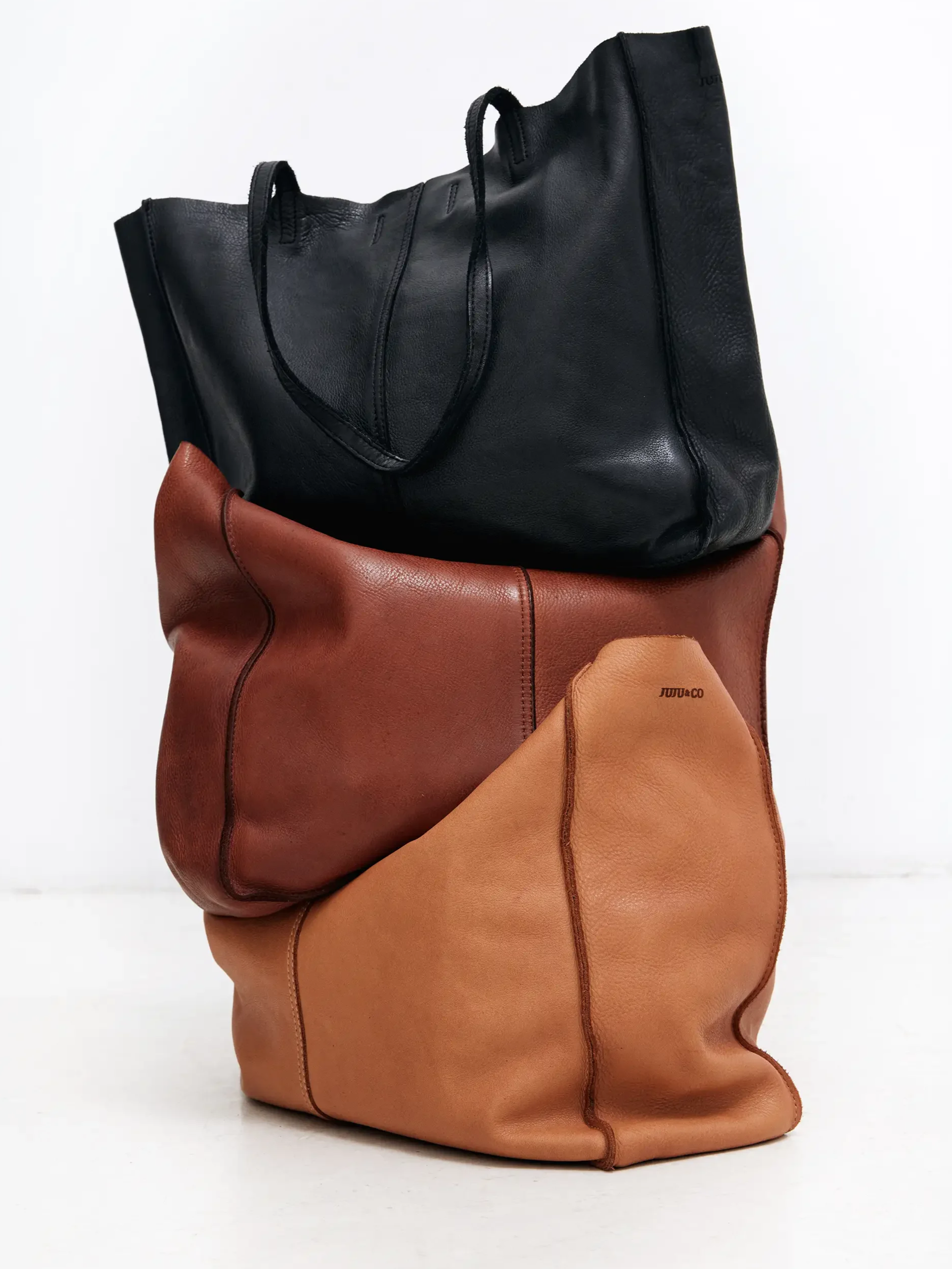 Three stacked leather tote bags in black, brown, and tan colors on a white background.