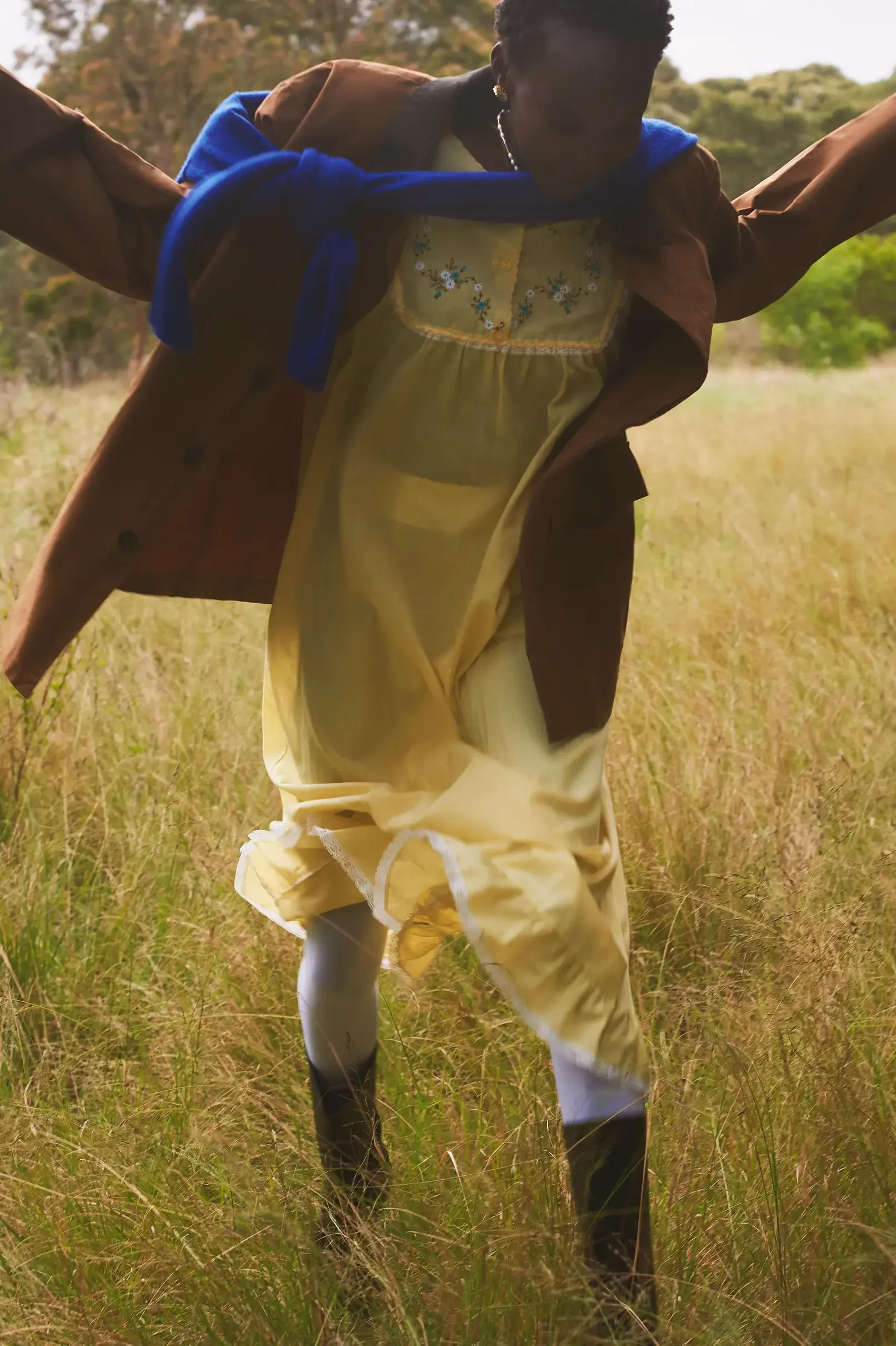 A person wearing yellow dress, brown jacket, white socks, black boots, and blue scarf standing in a grassy field with trees in the background.
