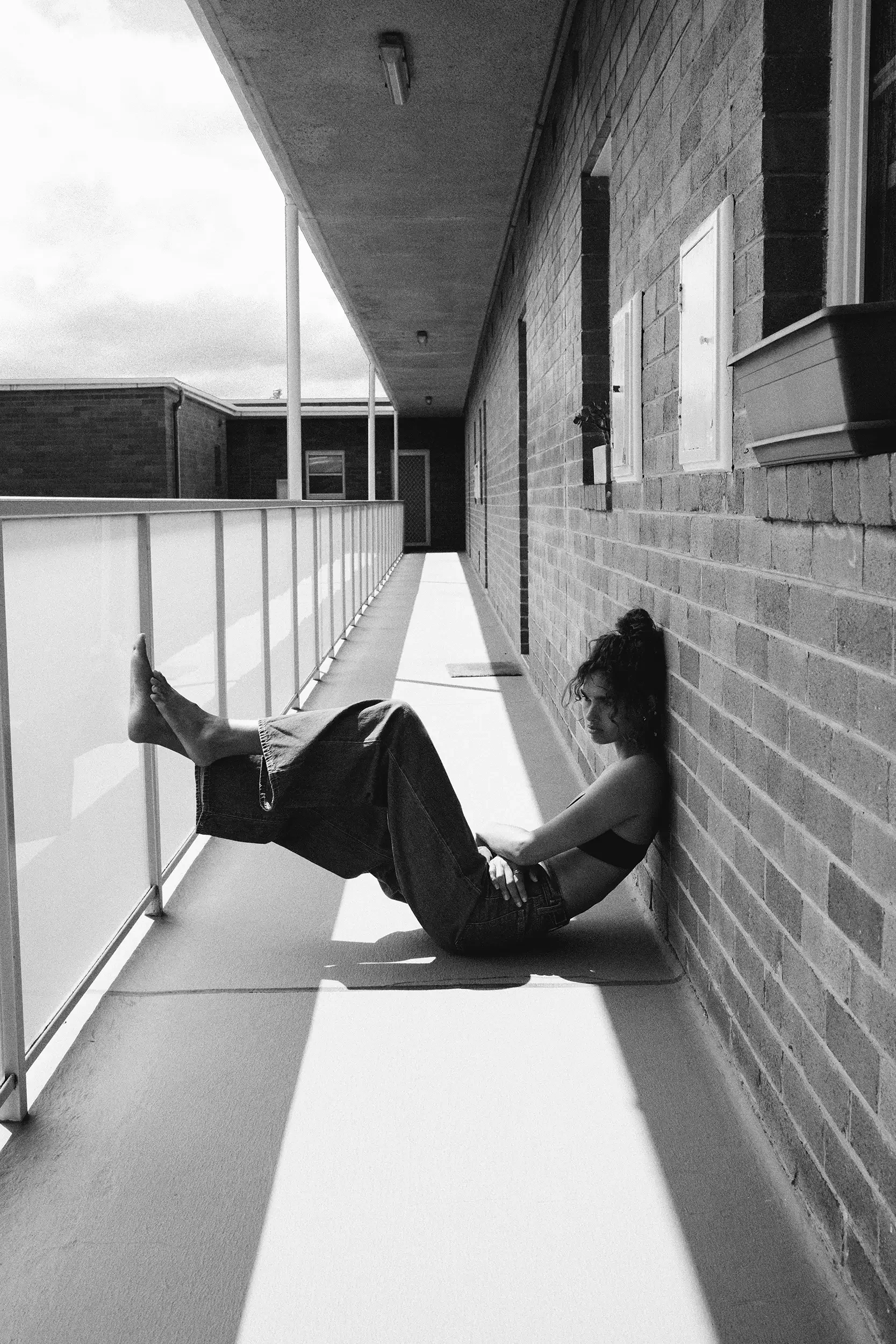 A woman with curly hair in a ponytail, wearing a black crop top and baggy jeans, sitting on a balcony floor against a brick wall, looking downward.