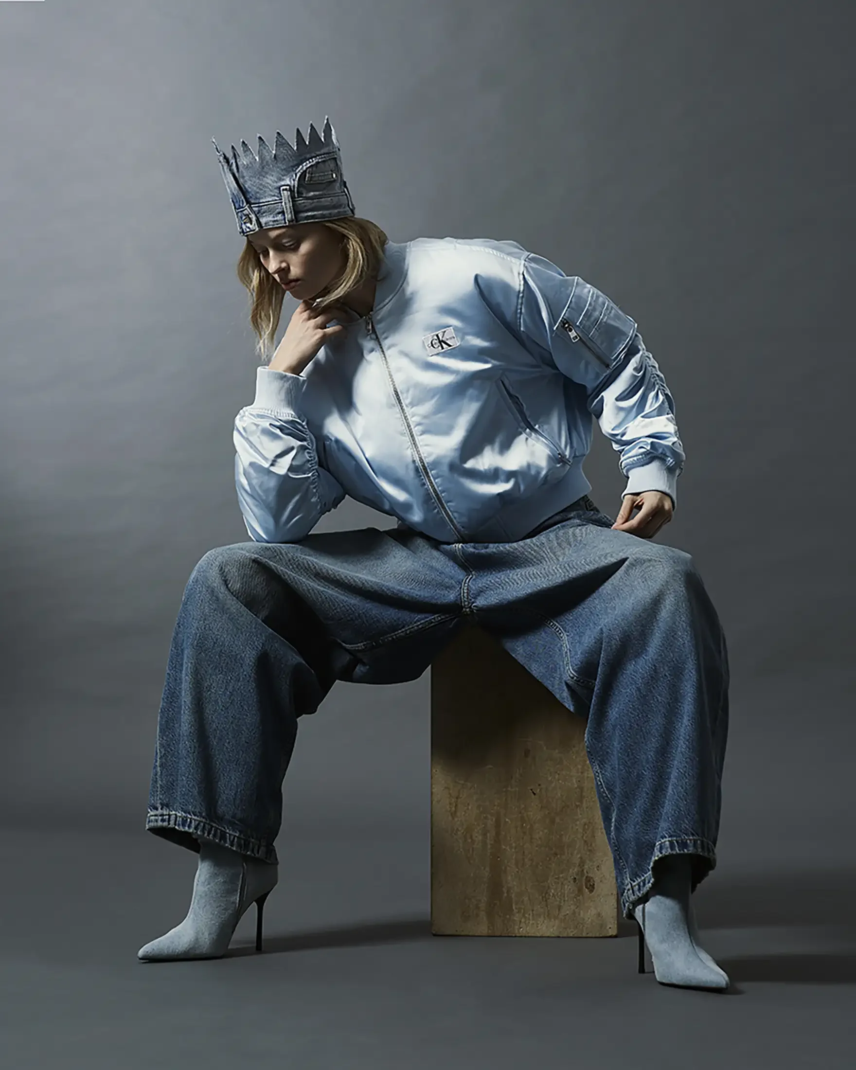 A woman wearing a denim crown, light blue bomber jacket, wide-leg jeans, and high-heeled boots sitting on a wooden block against a gray background.