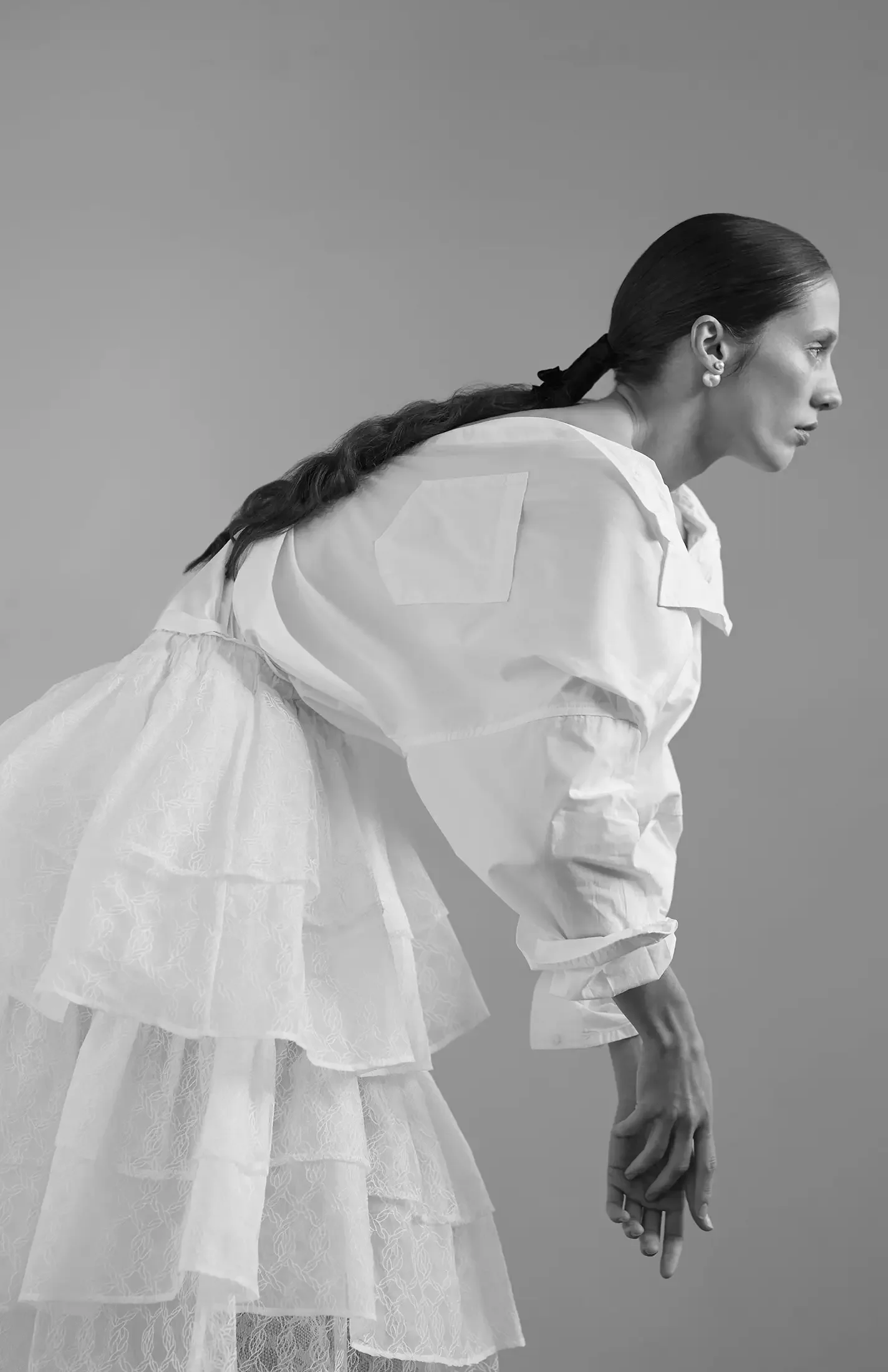 A black and white photo of a woman with long, dark hair in a ponytail, wearing a layered, sheer, lace skirt and an oversized white shirt. She is leaning forward slightly and looking to her right against a plain background.