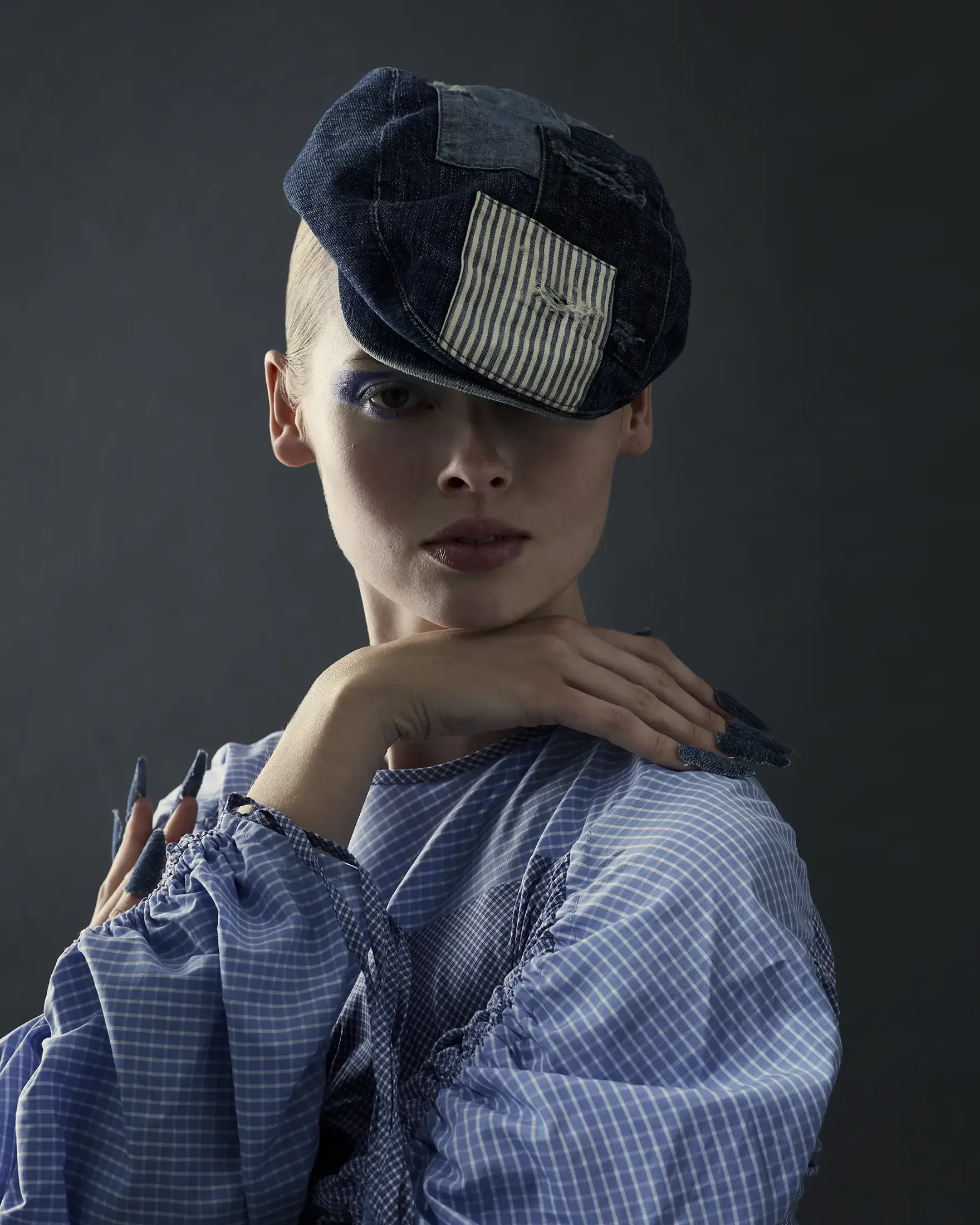 A fashion model wearing a denim hat with a striped patch and a blue checkered top, posing with her hand under her chin against a dark background.