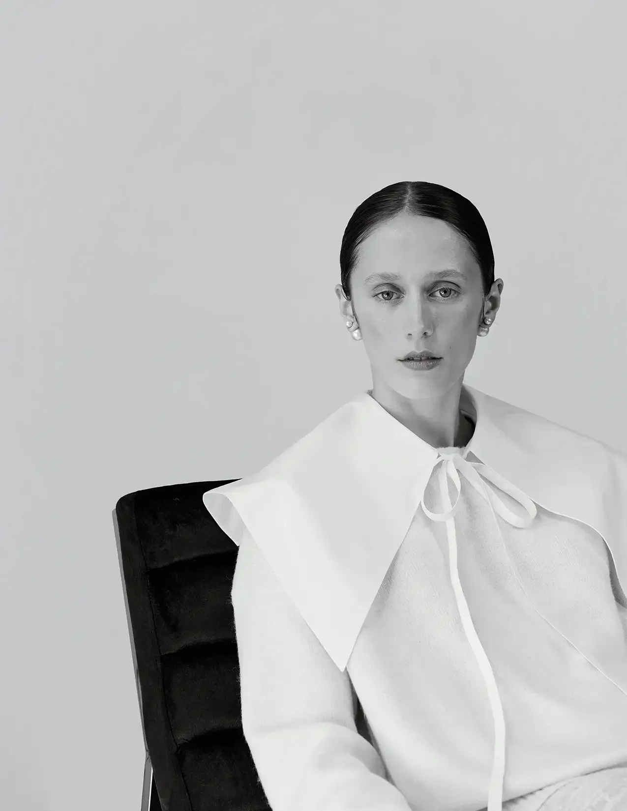 Black and white portrait of a woman with dark hair pulled back, wearing a white blouse with a large collar and bow, sitting on a black chair against a plain background.