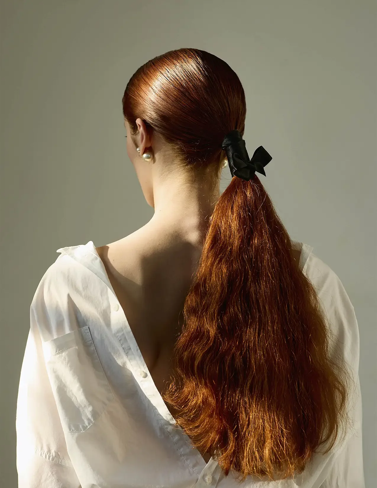 A woman with long, curly red hair tied back with a black ribbon, wearing a white shirt and pearl earrings, looking away from the camera.