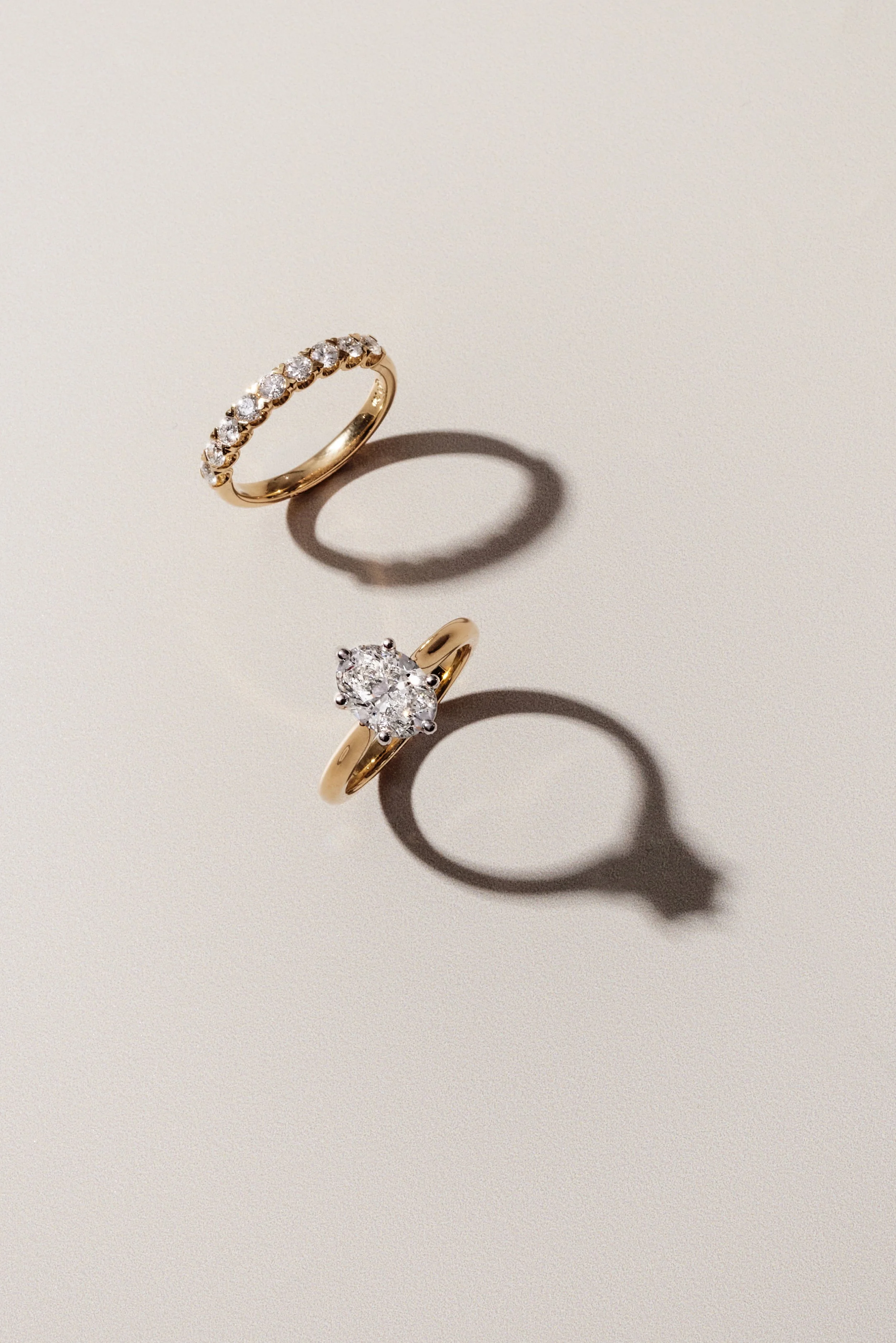 Two gold rings with diamonds, one with a row of small diamonds and the other with a single large diamond, placed on a light-colored surface with shadows.