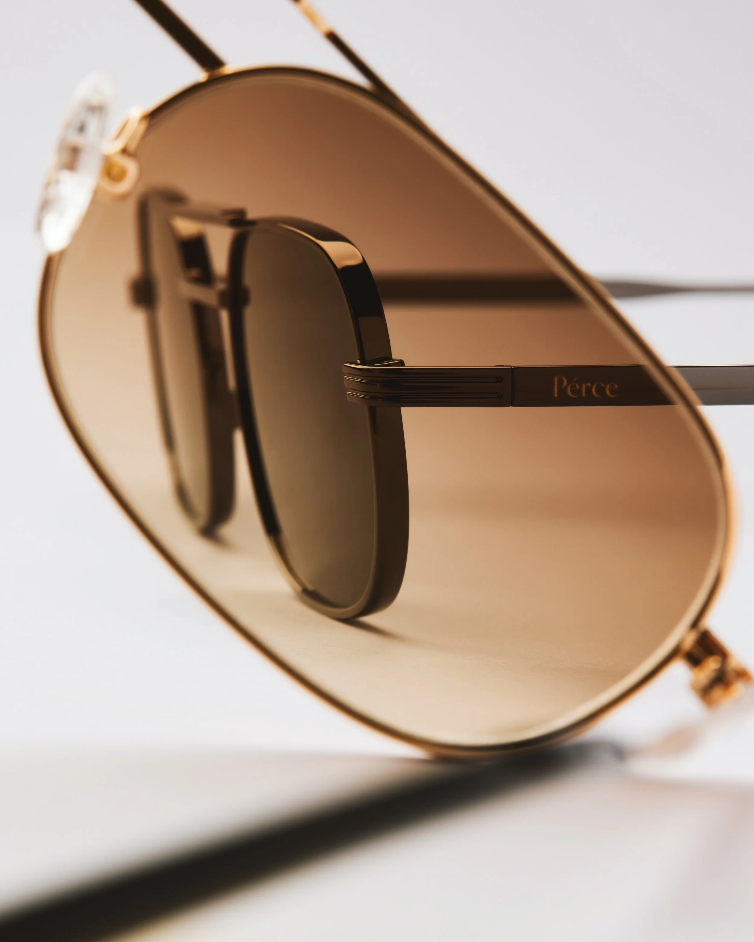 Close-up of a gold and black sunglass with the brand name 'Pérce' on the arm, with a blurred background.