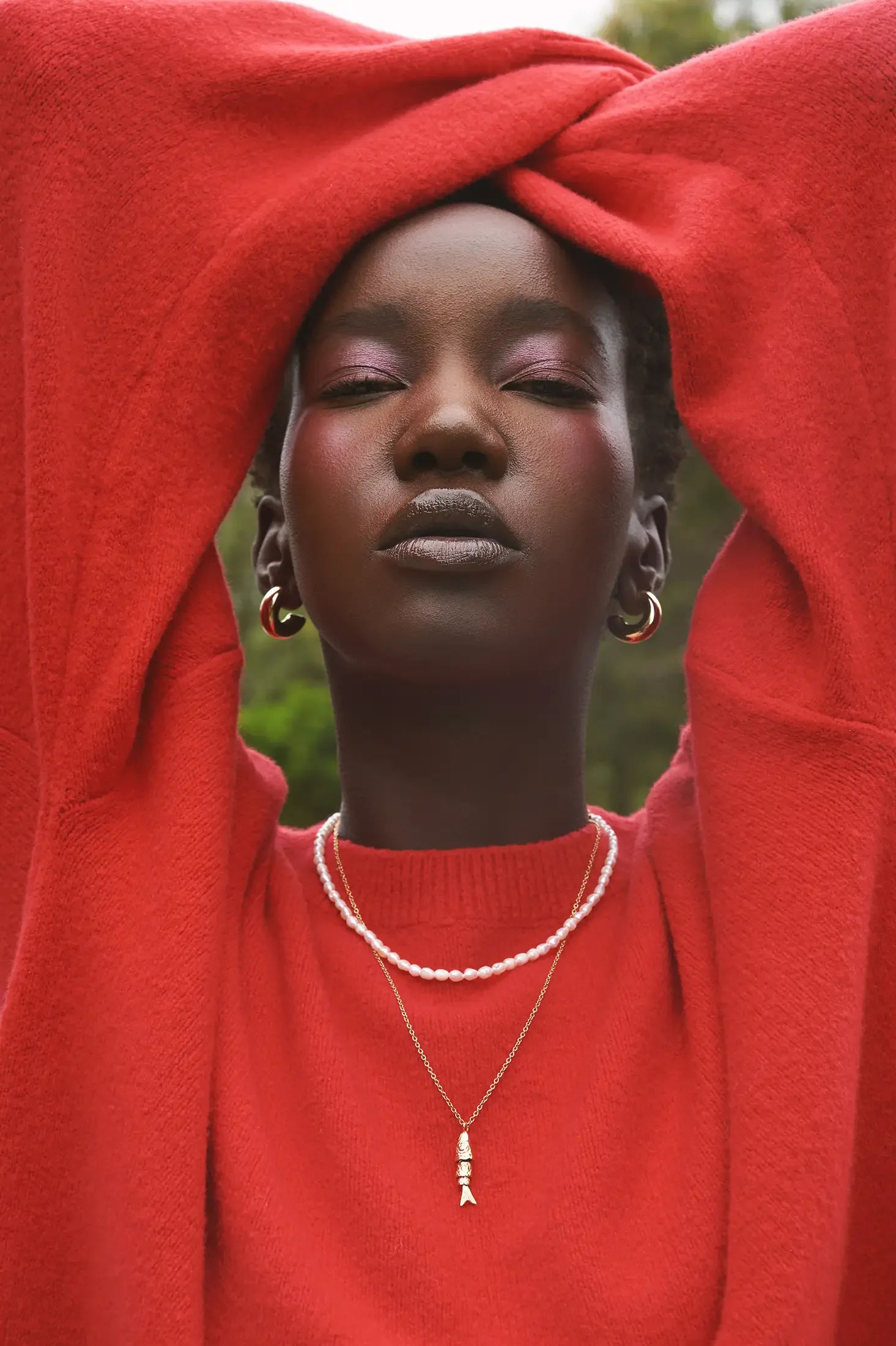 A young Black woman with short hair, wearing a red fleece hoodie, layered necklaces, and hoop earrings, posing outdoors with a blurred green background.