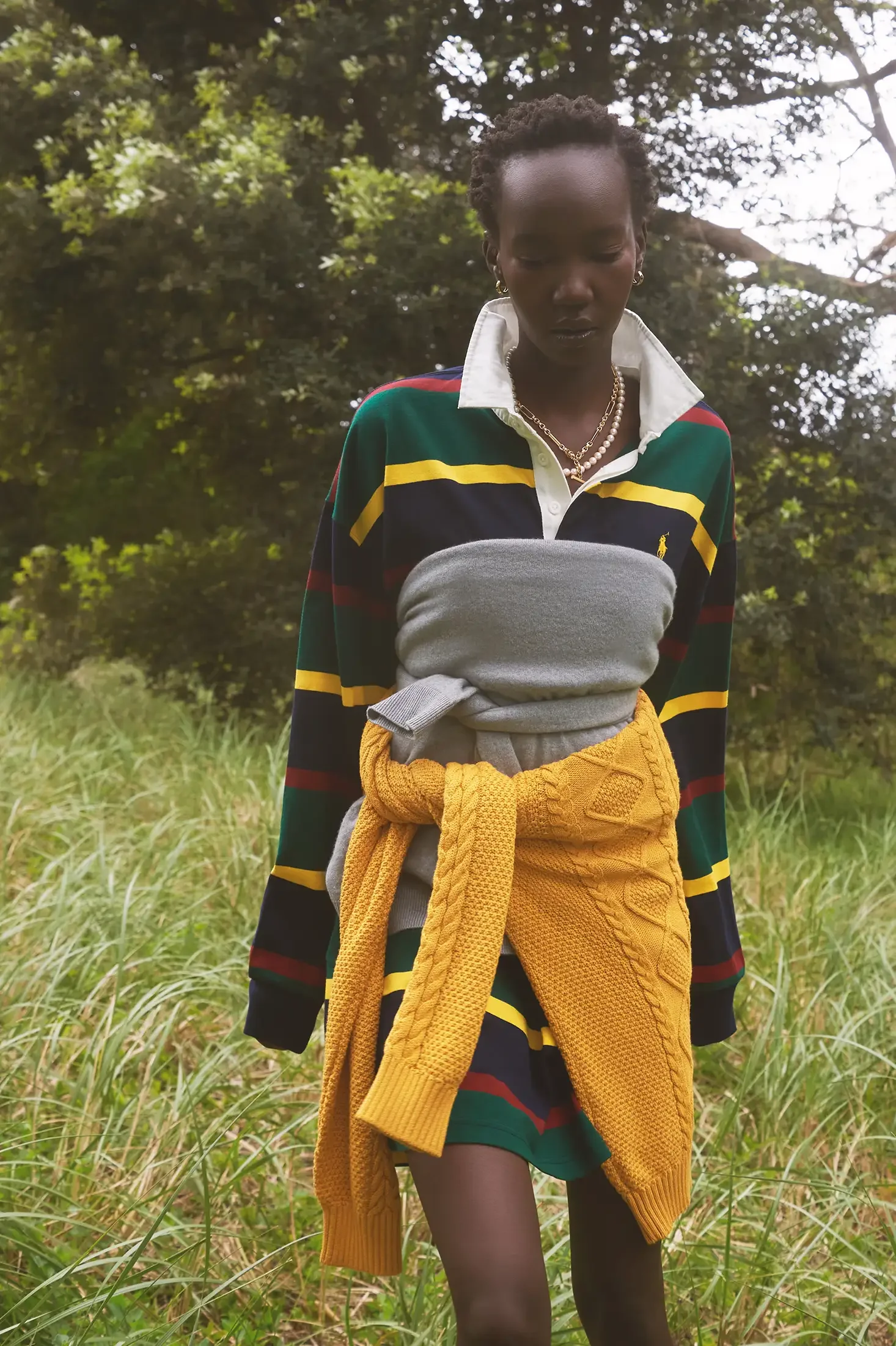 A woman standing outdoors in a grassy area wearing a striped rugby shirt with a yellow cardigan tied around her waist and layered necklaces.