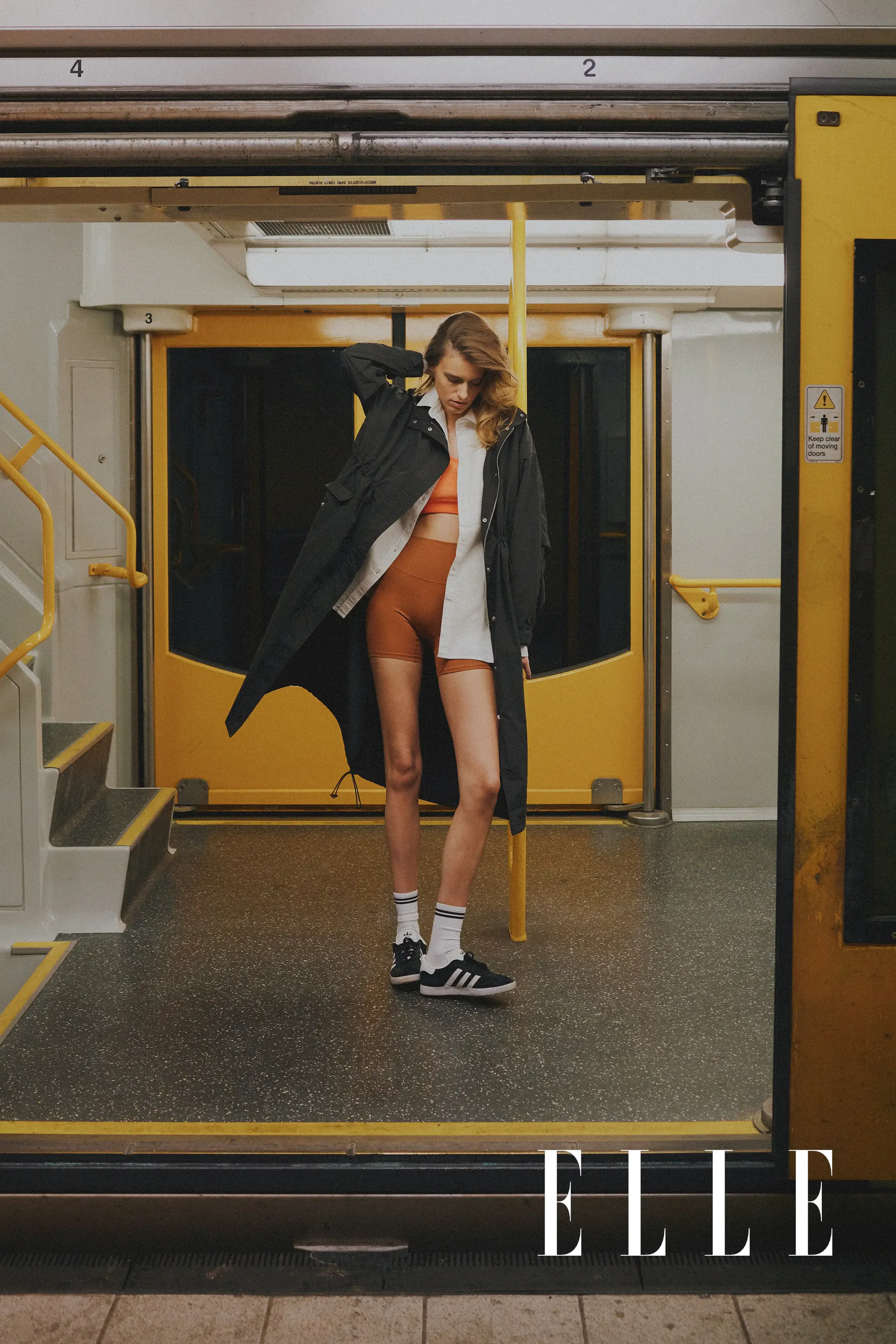 A woman in athletic wear, including a black jacket, orange sports bra, matching shorts, white socks with black stripes, and sneakers, stands in a subway entrance, adjusting her hair. The background features yellow subway doors, a black doorway, and a