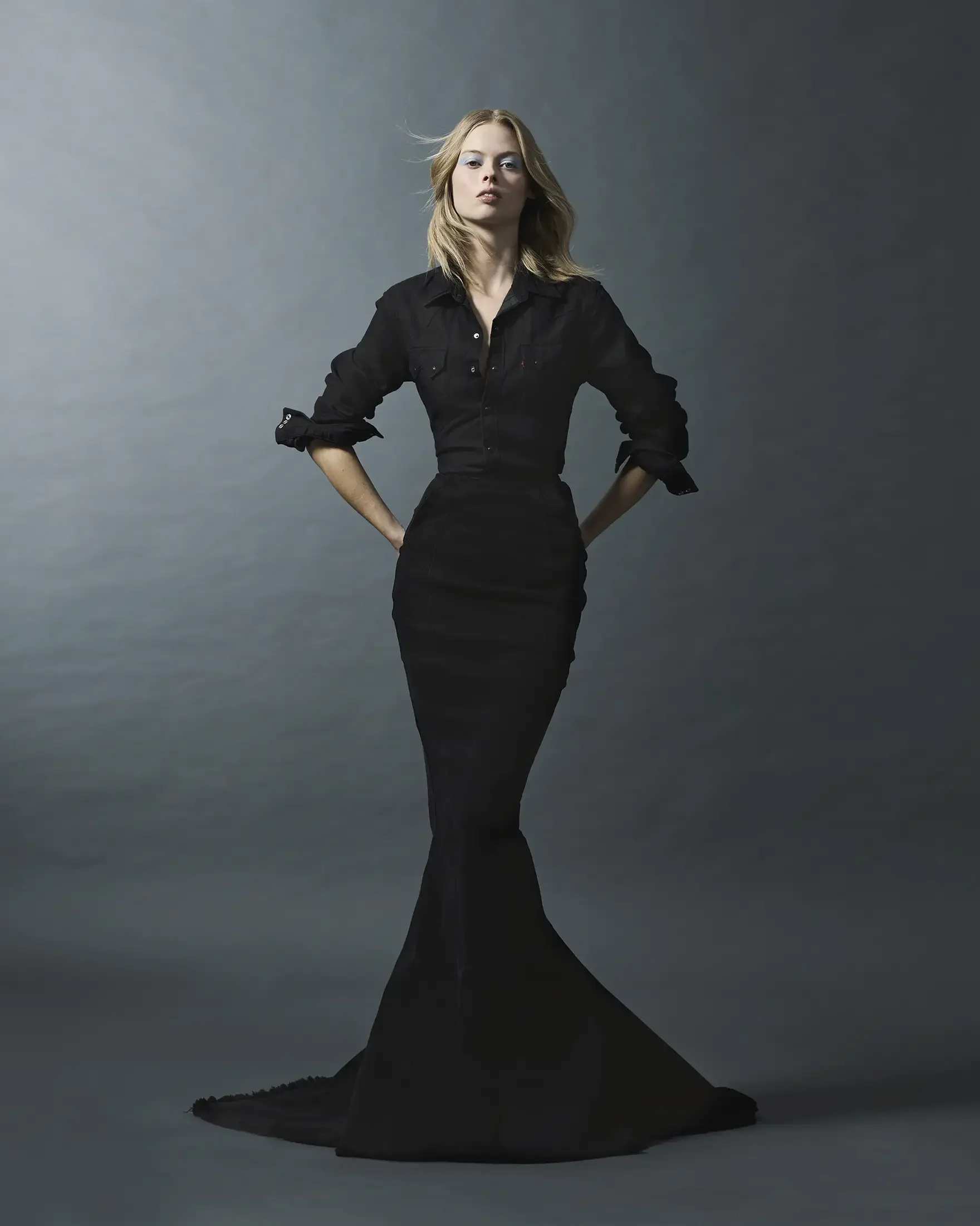 A woman in a black dress standing with hands on hips against a plain gray background.
