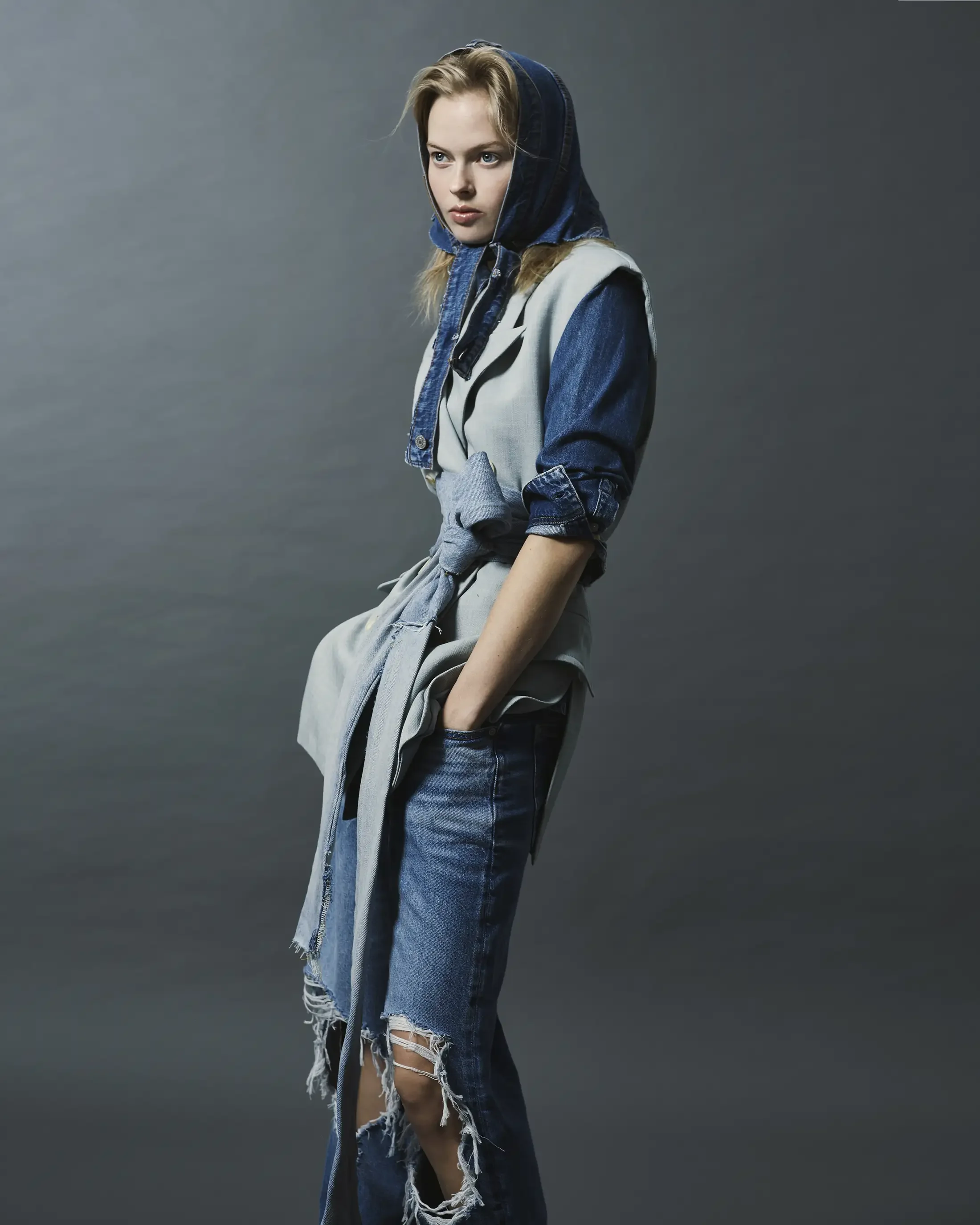 A young woman with blonde hair wearing a hooded denim jacket layered over a light gray vest, with a gray sweatshirt tied around her waist, and ripped jeans, standing against a plain gray background.