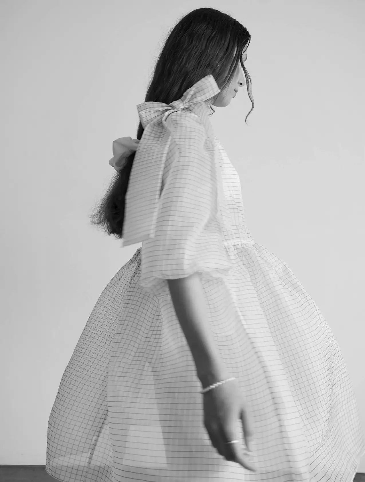 A black-and-white photo of a woman in profile, wearing a checkered dress with puffy sleeves, a large bow in her hair, and a bracelet on her wrist.