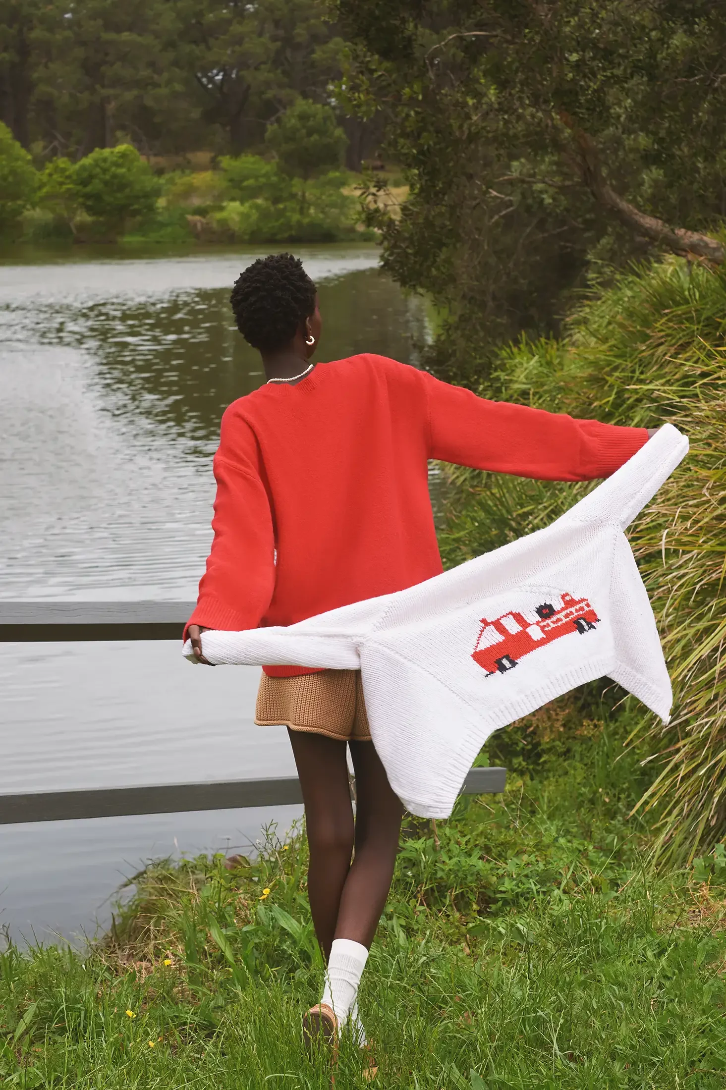 Person standing by a lake, holding a large white sock with a red house design, dressed in a red sweater and tan skirt, with black tights and white socks.