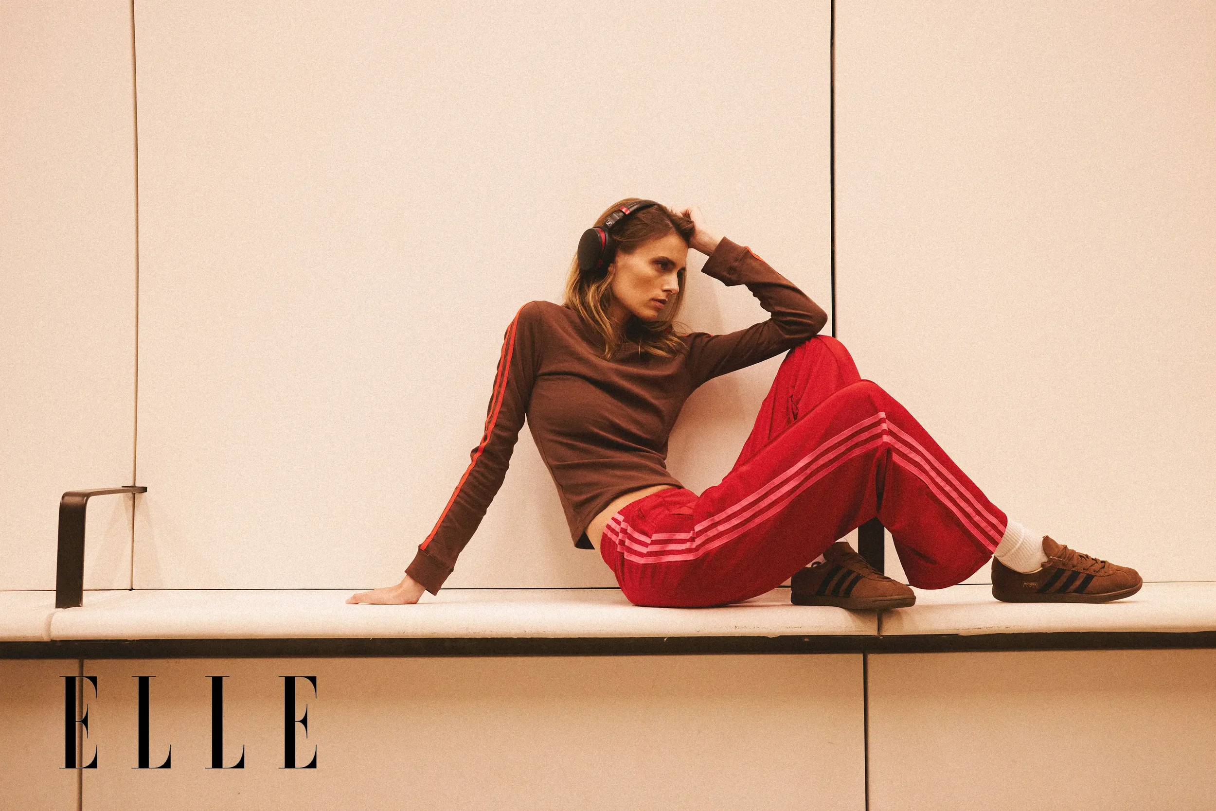 A woman with long brown hair dressed in a brown and red athletic outfit, sitting on a bench against a plain wall, wearing headphones, with one hand on her head and the other on the bench, looking thoughtful or tired.