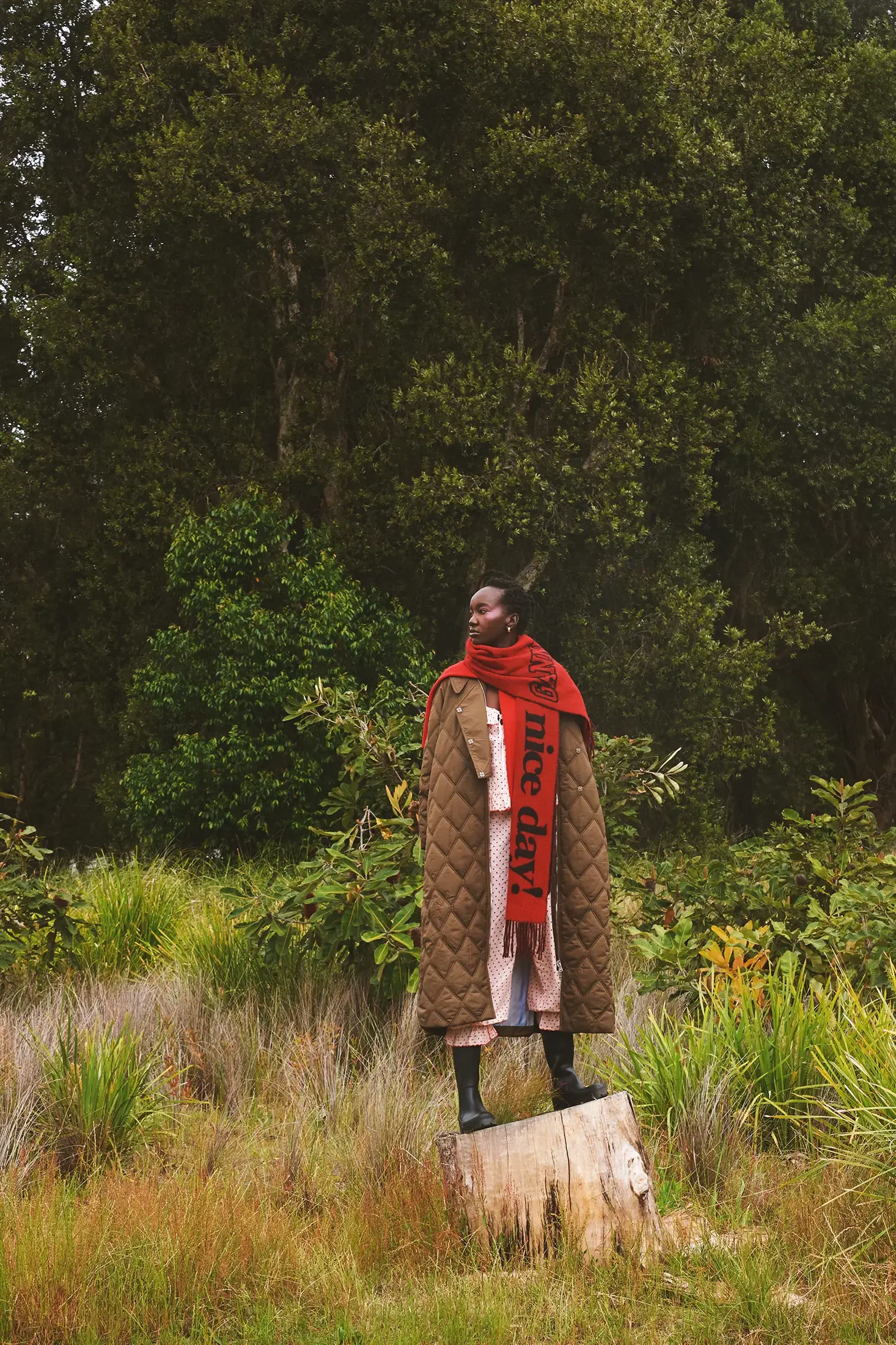 A woman standing on a tree stump in a grassy clearing with dense green trees in the background. She is wearing a long brown quilted coat, pink pants, black boots, and a bright red scarf with black text.