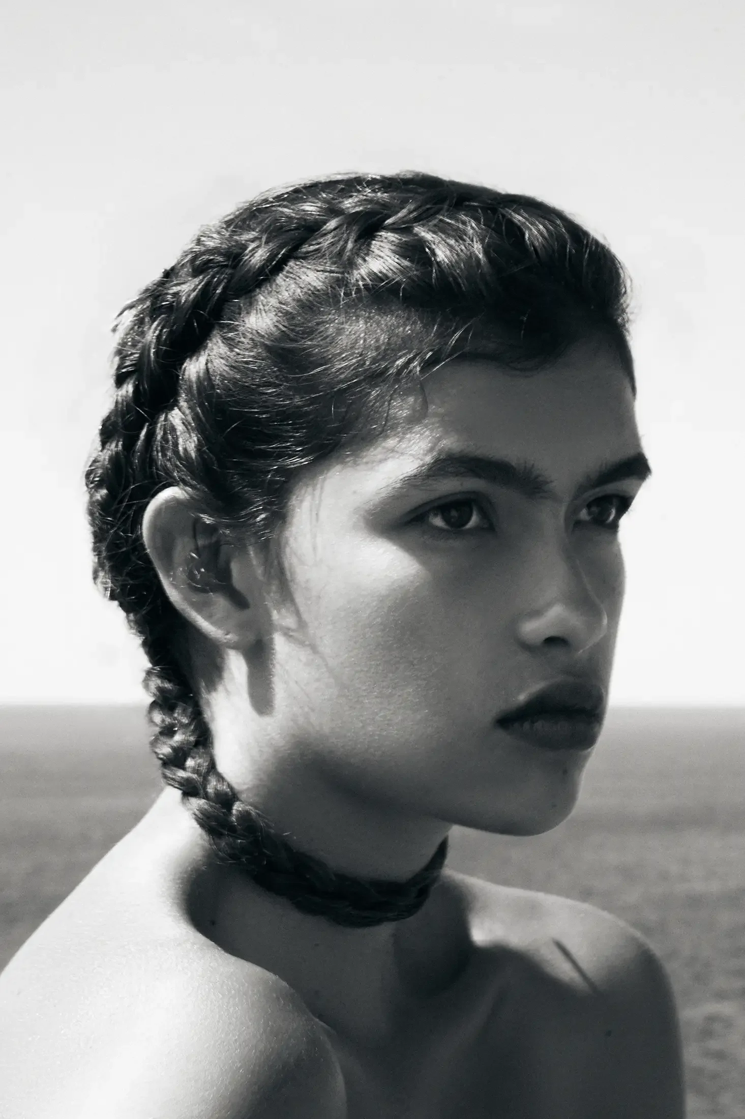 A black and white photo of a woman with braided hair and a choker, gazing off into the distance outdoors.