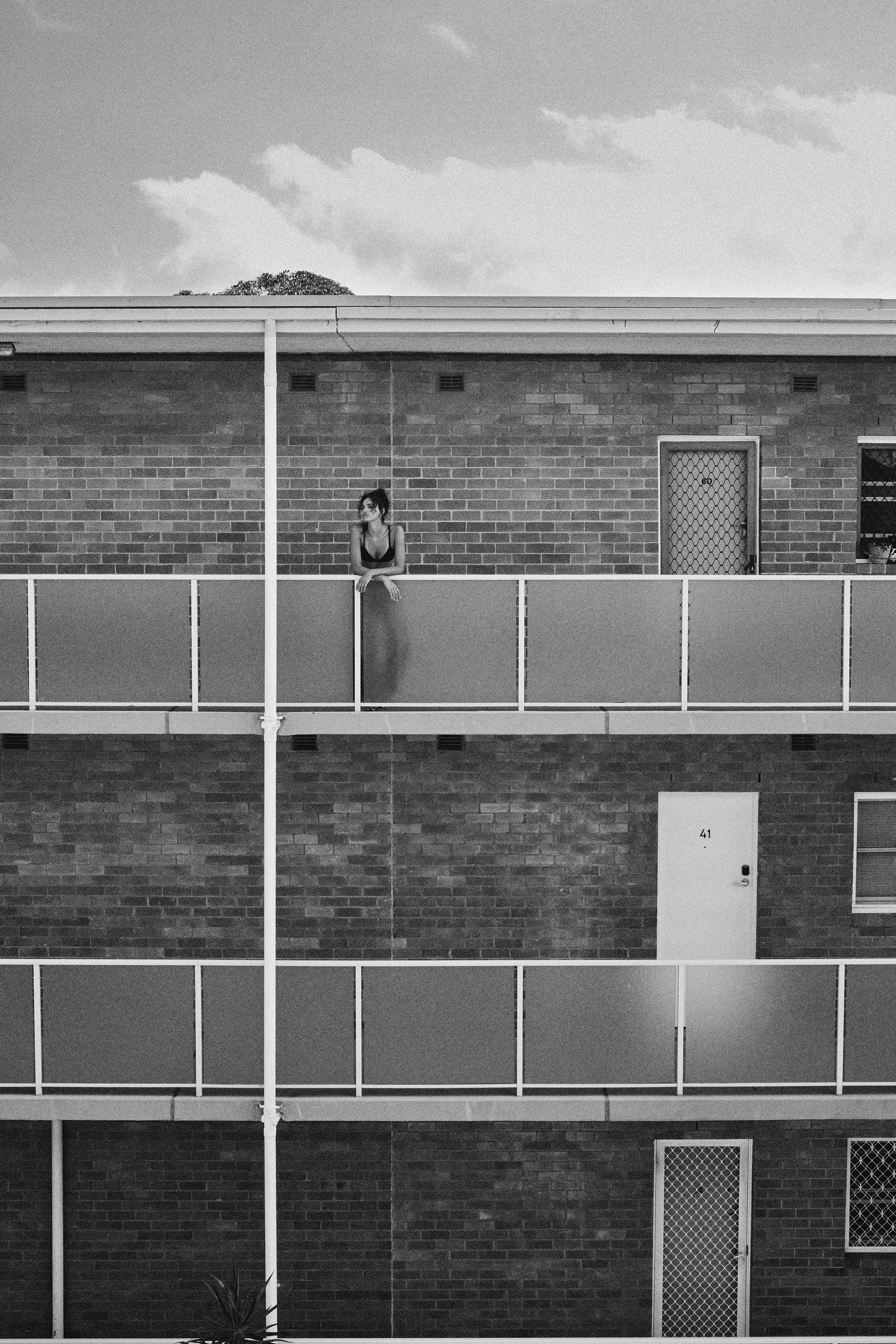 A woman standing on a balcony of an apartment building, looking to the side, with brick walls and doors numbered 41 and 60 behind her, in black and white.