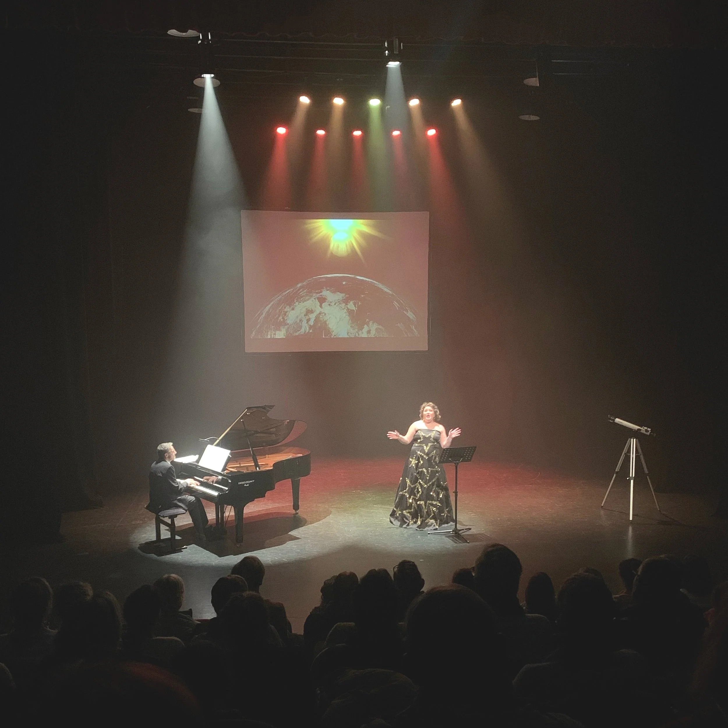 Une performance sur scène avec un chanteur et un pianiste, une projection d'une image de la Terre avec le soleil, et un télescope sur un trépied, sous des projecteurs colorés.